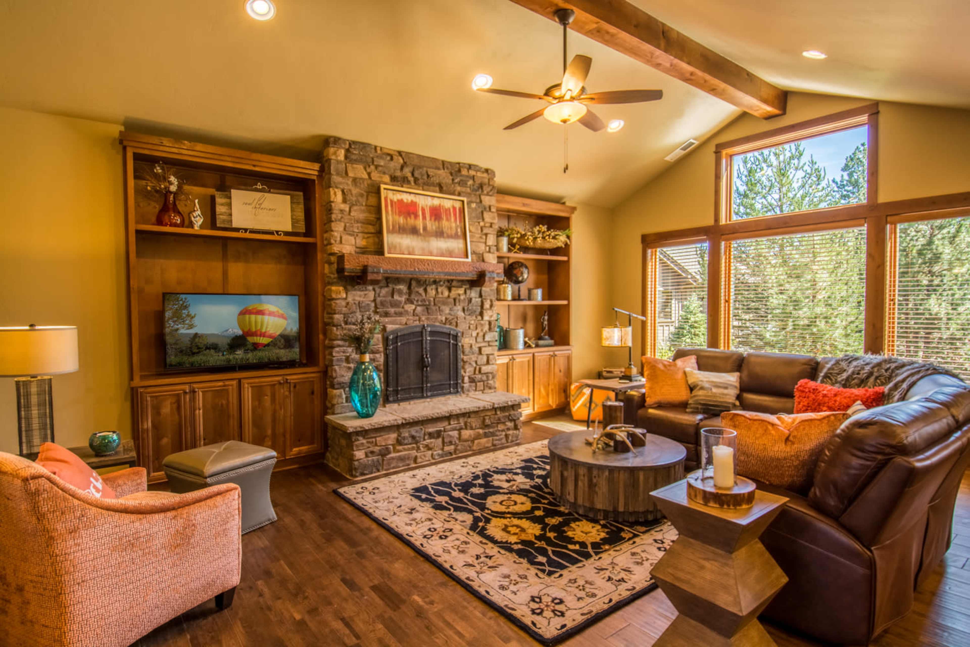 The image shows a cozy living room with a stone fireplace, wooden shelves, a large round coffee table, and ample natural light coming through large windows.