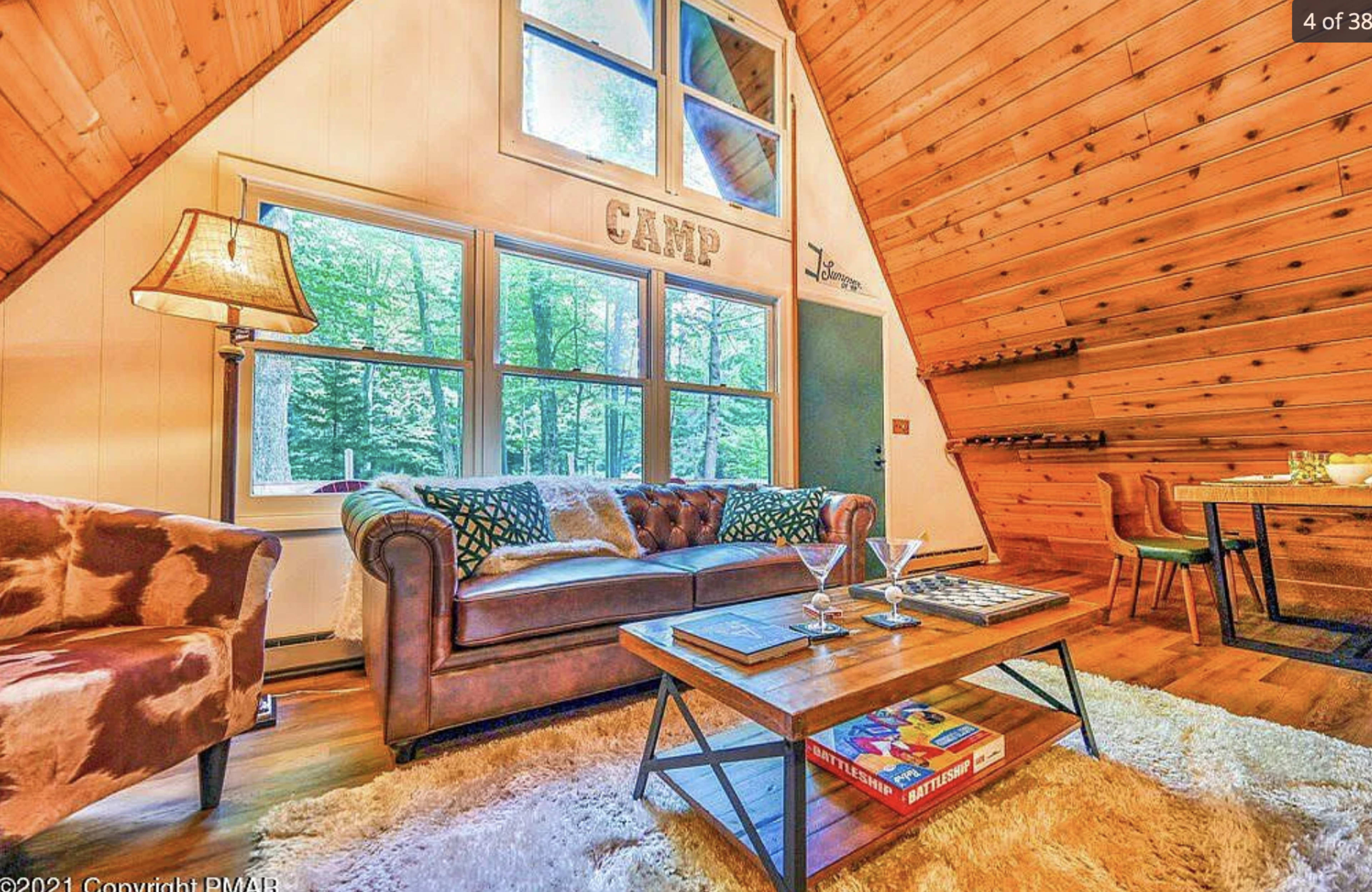 The interior features a cozy living area with a leather sofa, a wooden coffee table, and large windows overlooking a forested view.