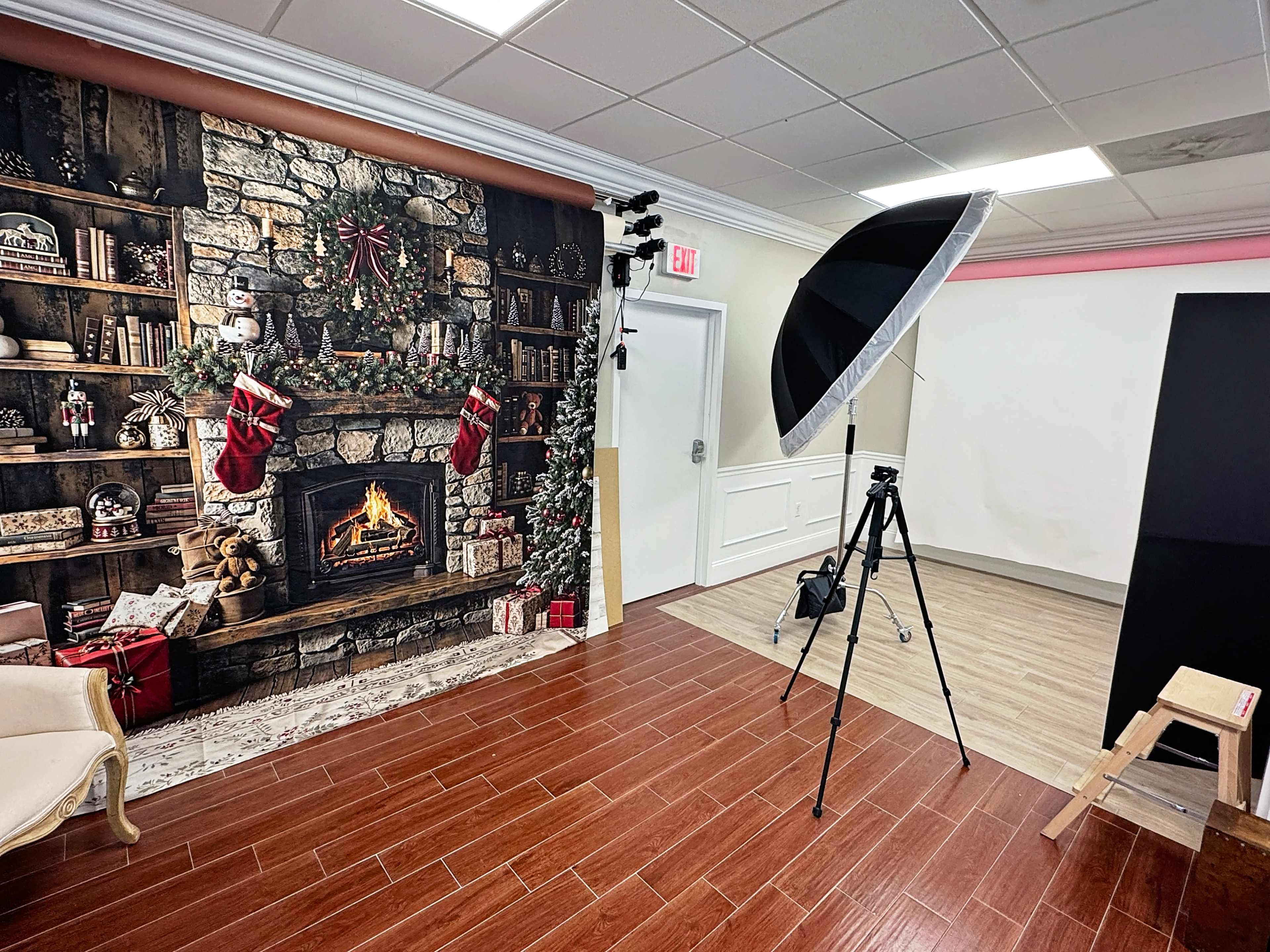 The scene features a photography studio with a decorated backdrop resembling a cozy fireplace surrounded by bookshelves and holiday stockings, along with a large lighting umbrella and a tripod.