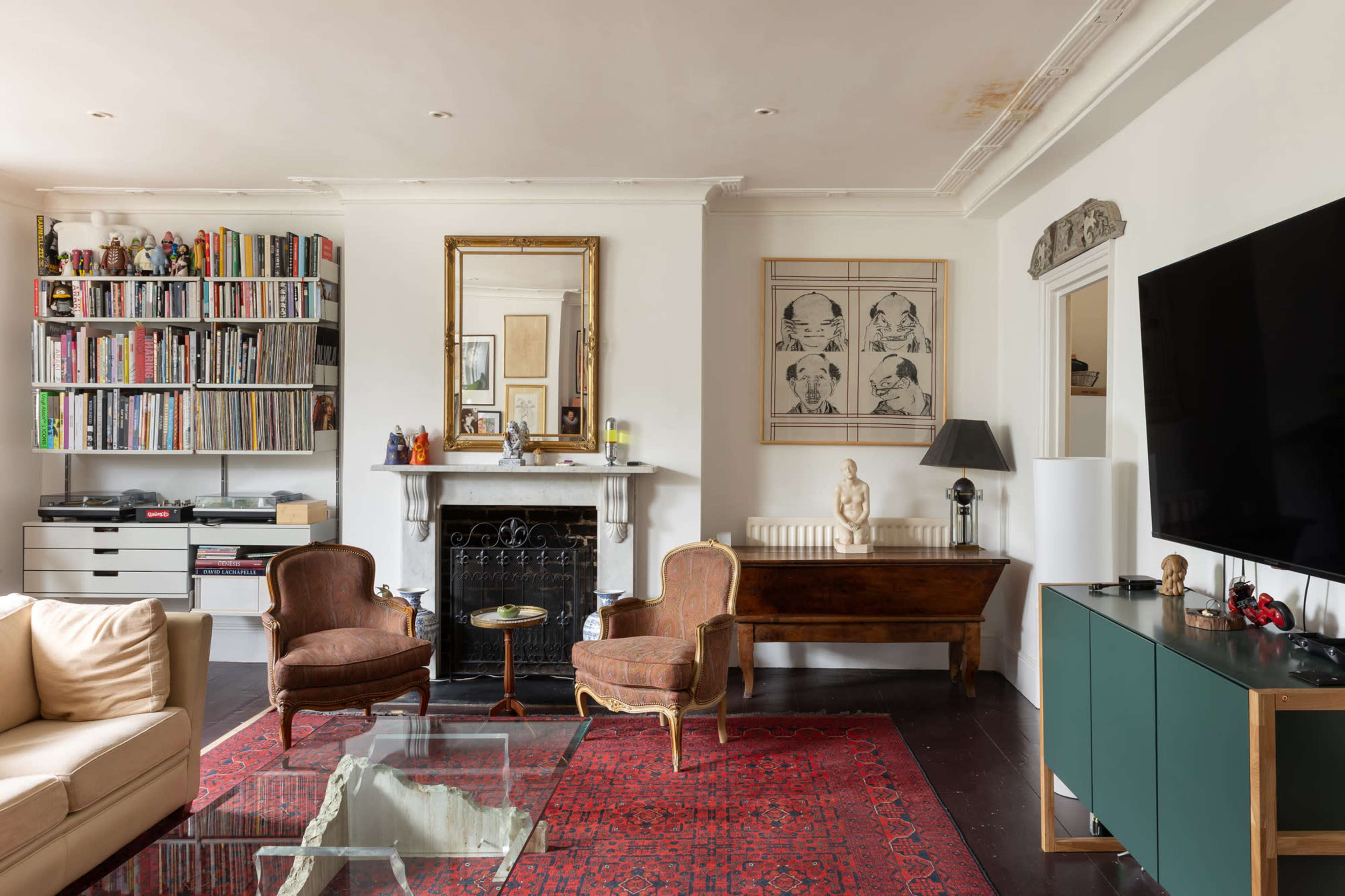 A living room features a pair of vintage armchairs, a fireplace with decorative art, a large television, and shelves filled with books against a light-colored wall.