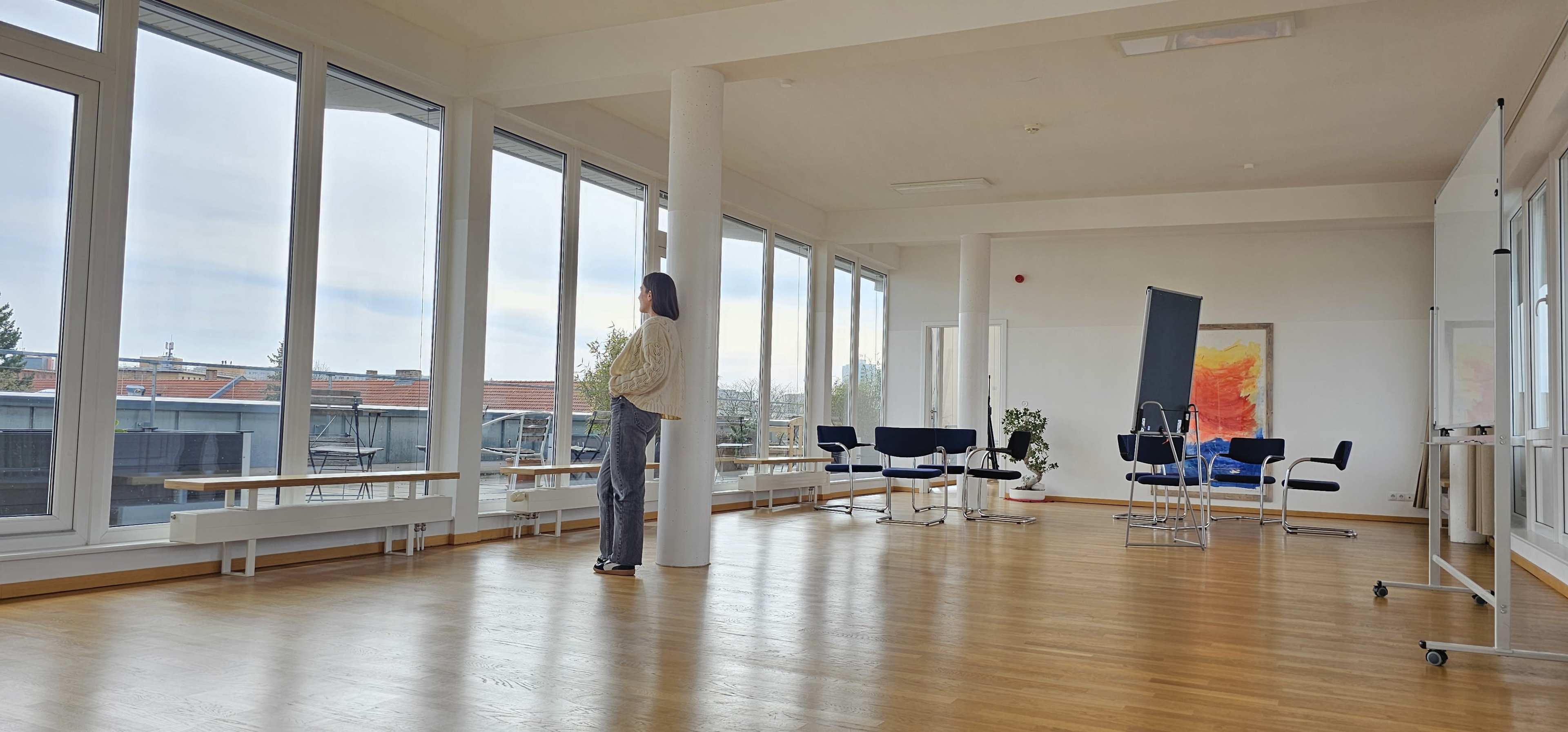 A person stands near large windows in a bright, spacious room with wooden floors and modern furniture.