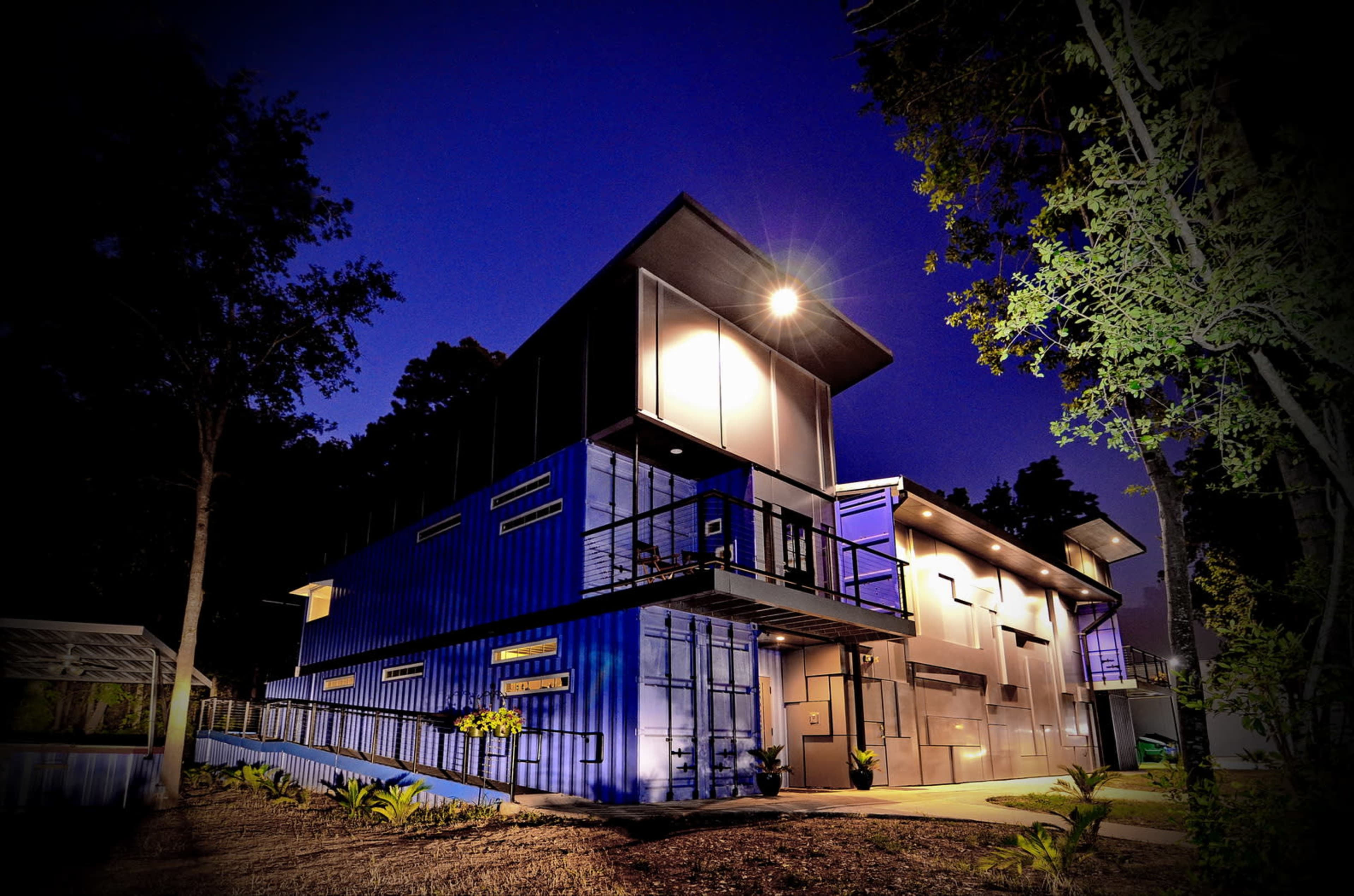 The image shows a modern building made from shipping containers, illuminated at night and surrounded by trees.