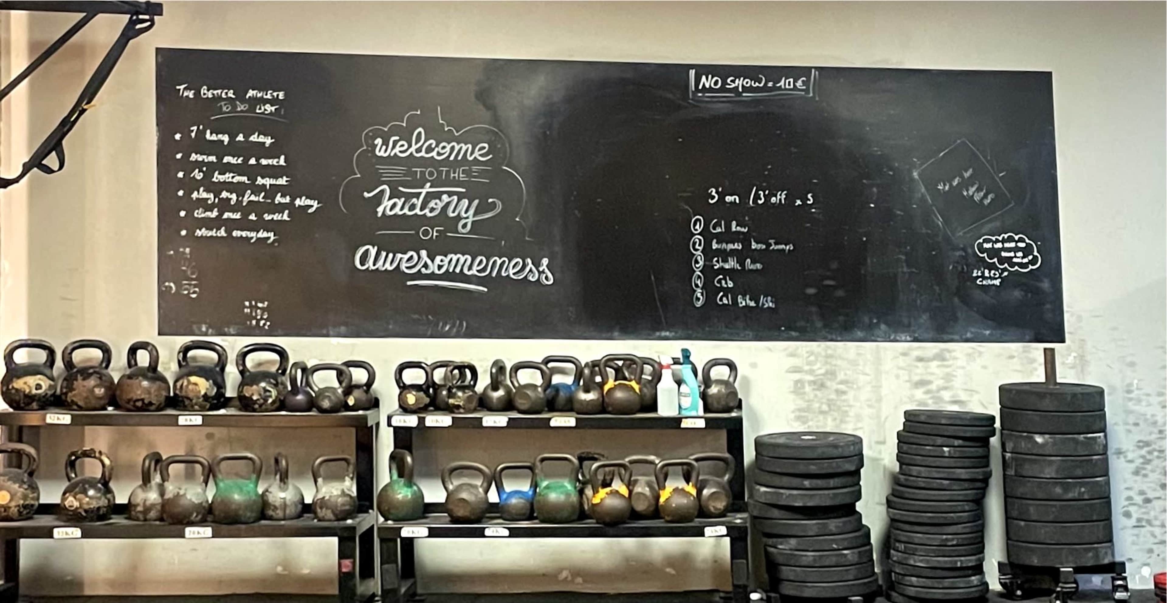 A gym space with a chalkboard displaying a welcome message and gym rules, alongside rows of kettlebells and weight plates on shelves.