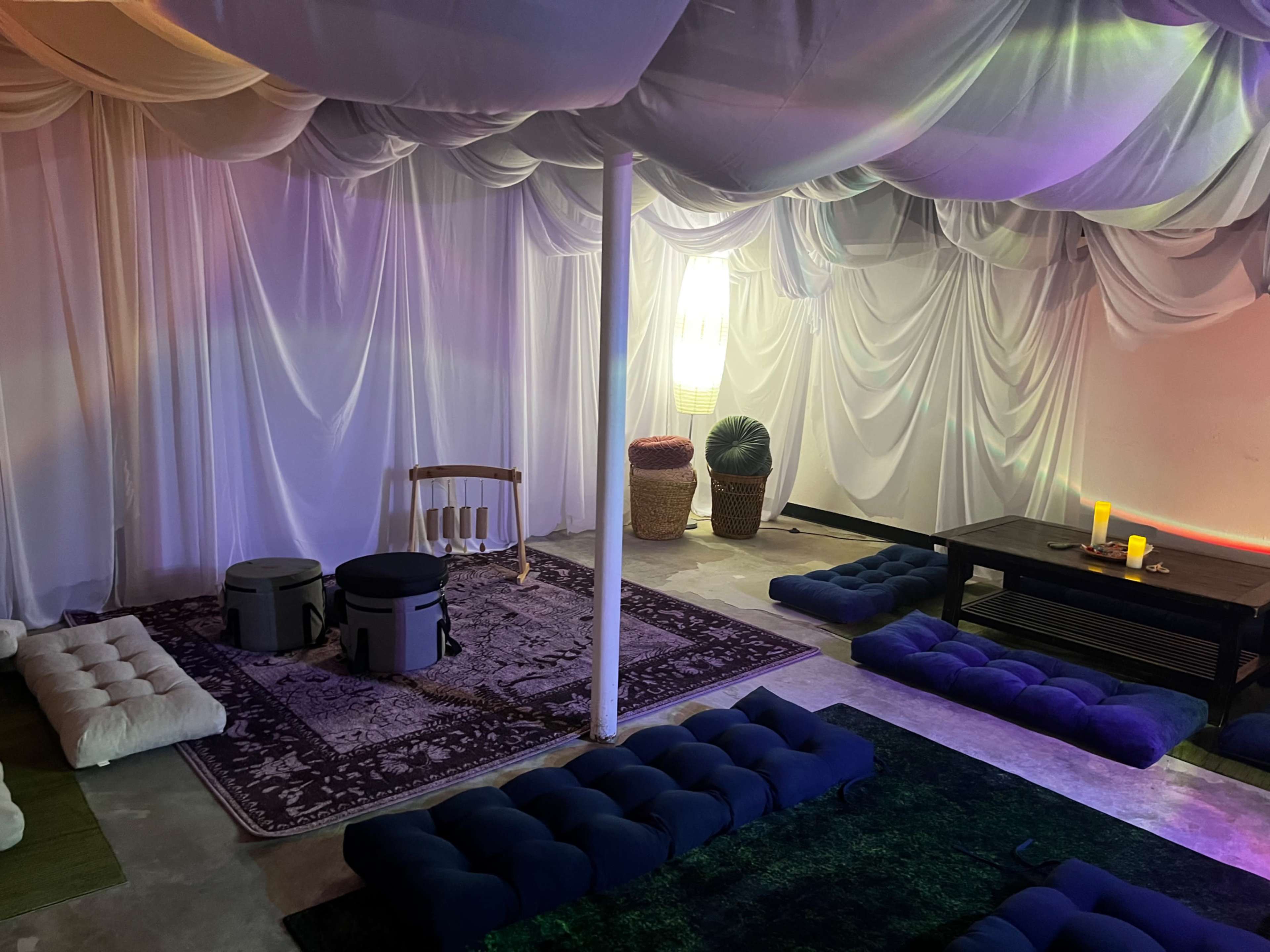 The scene shows a softly lit room with draped white fabric overhead, featuring cushions and mats arranged on the floor, and a low table with candles.