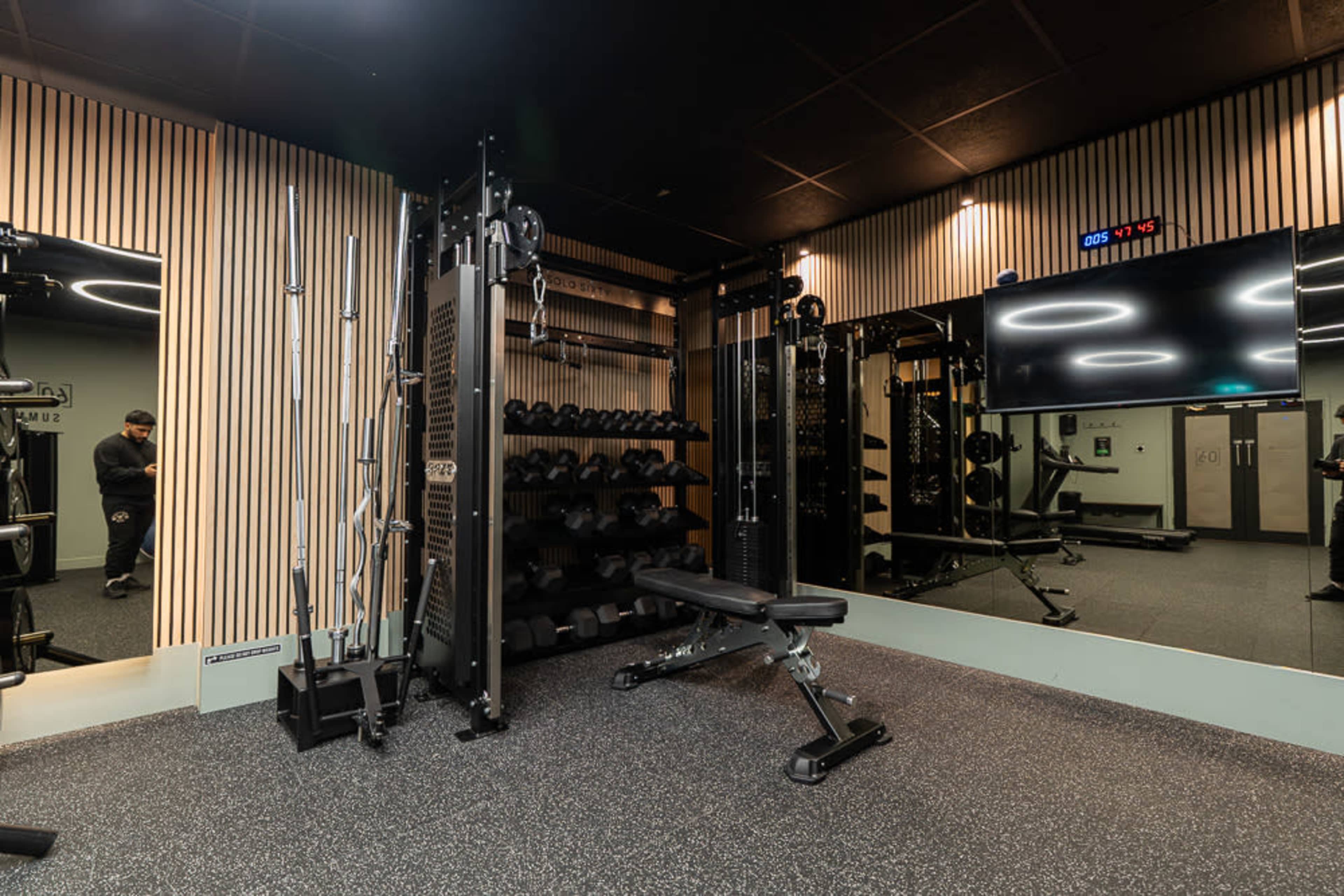 The image shows a modern gym space featuring a wall of free weights, a cable machine, and a bench, all set against a backdrop of wood paneling and a large wall-mounted screen.
