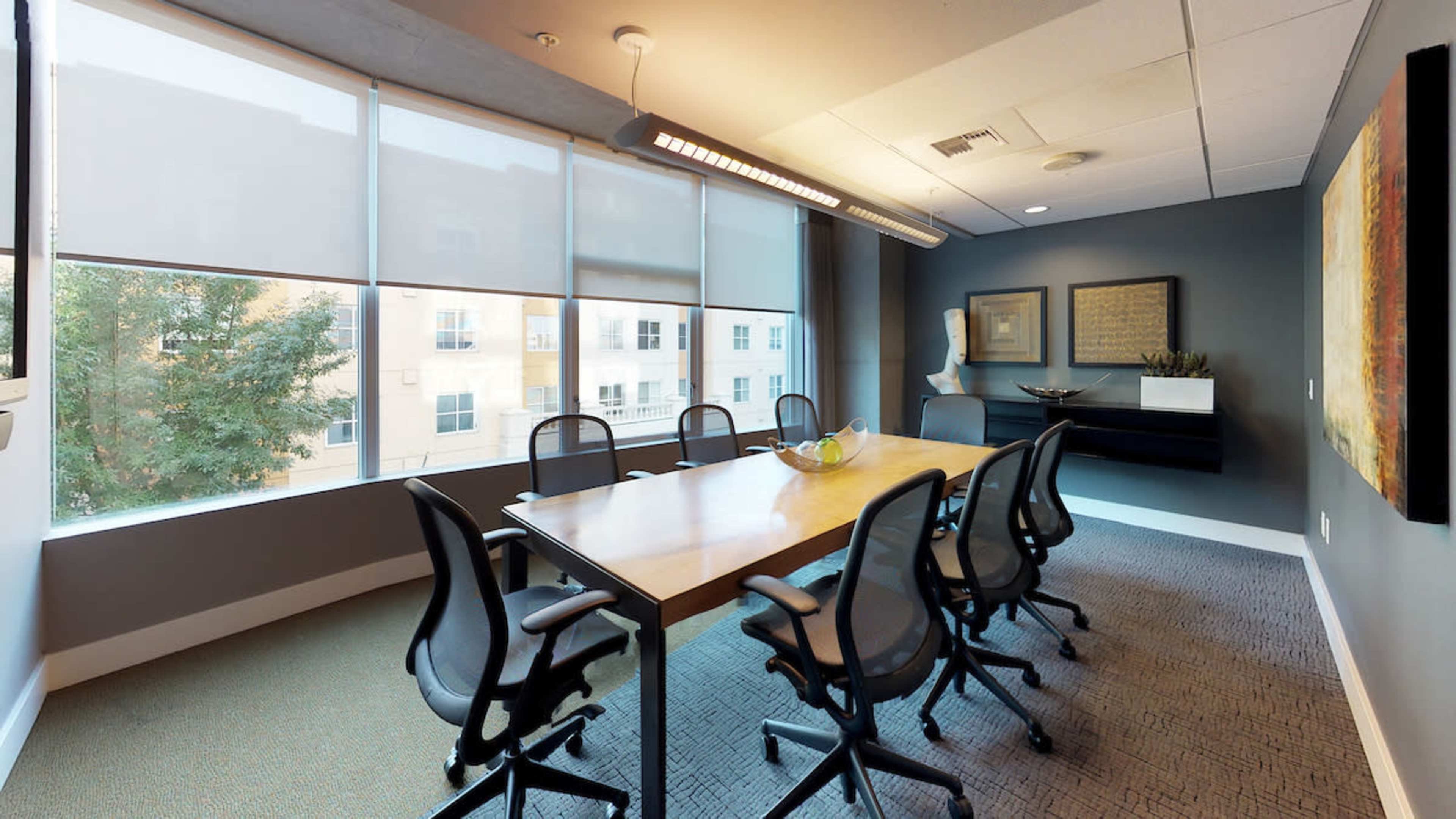 A modern conference room with a long wooden table surrounded by six office chairs and large windows letting in natural light.