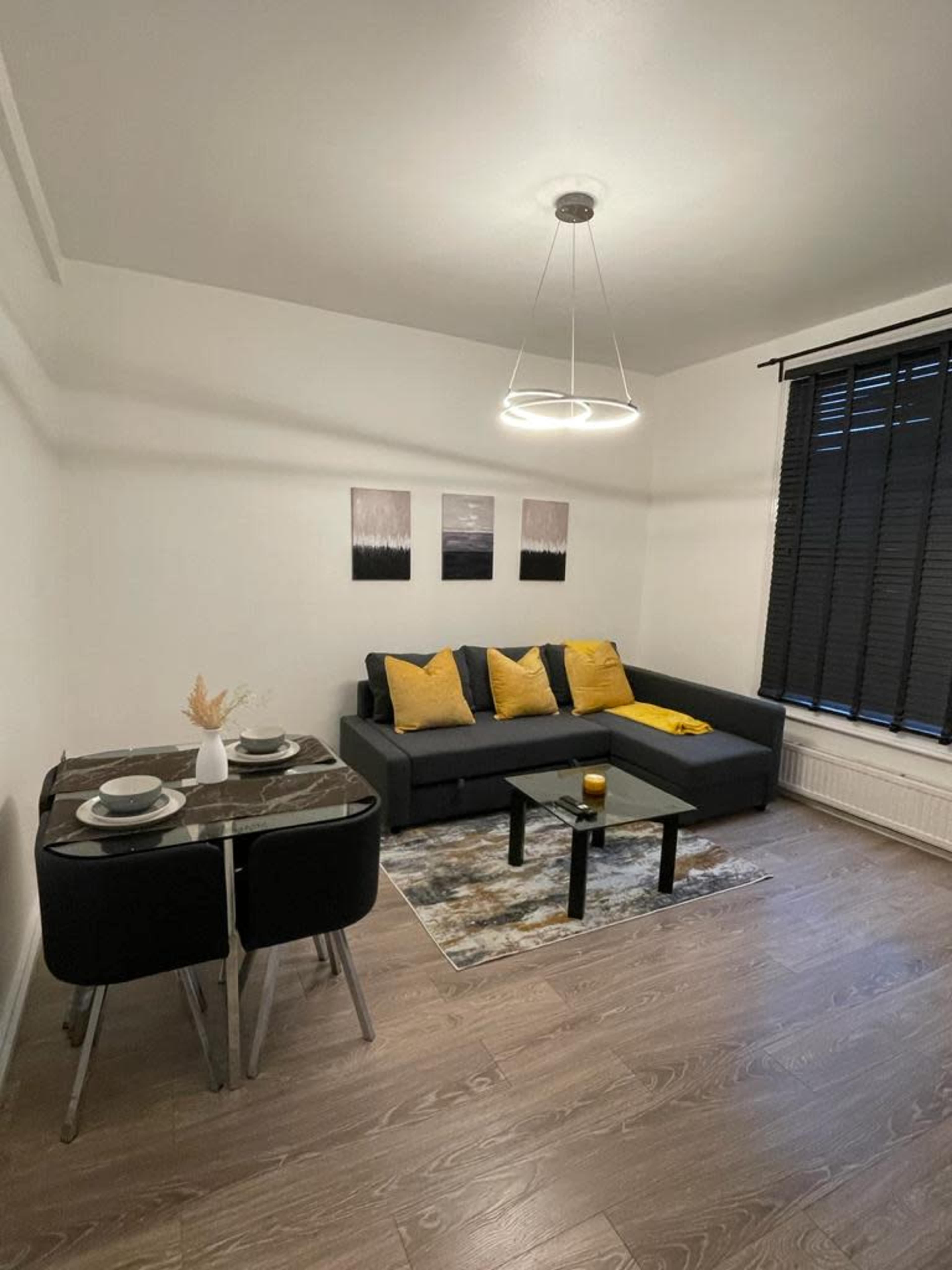 A cozy living space with a dark sectional sofa, two yellow accent pillows, a dining table set for two, and a rug on a wooden floor.