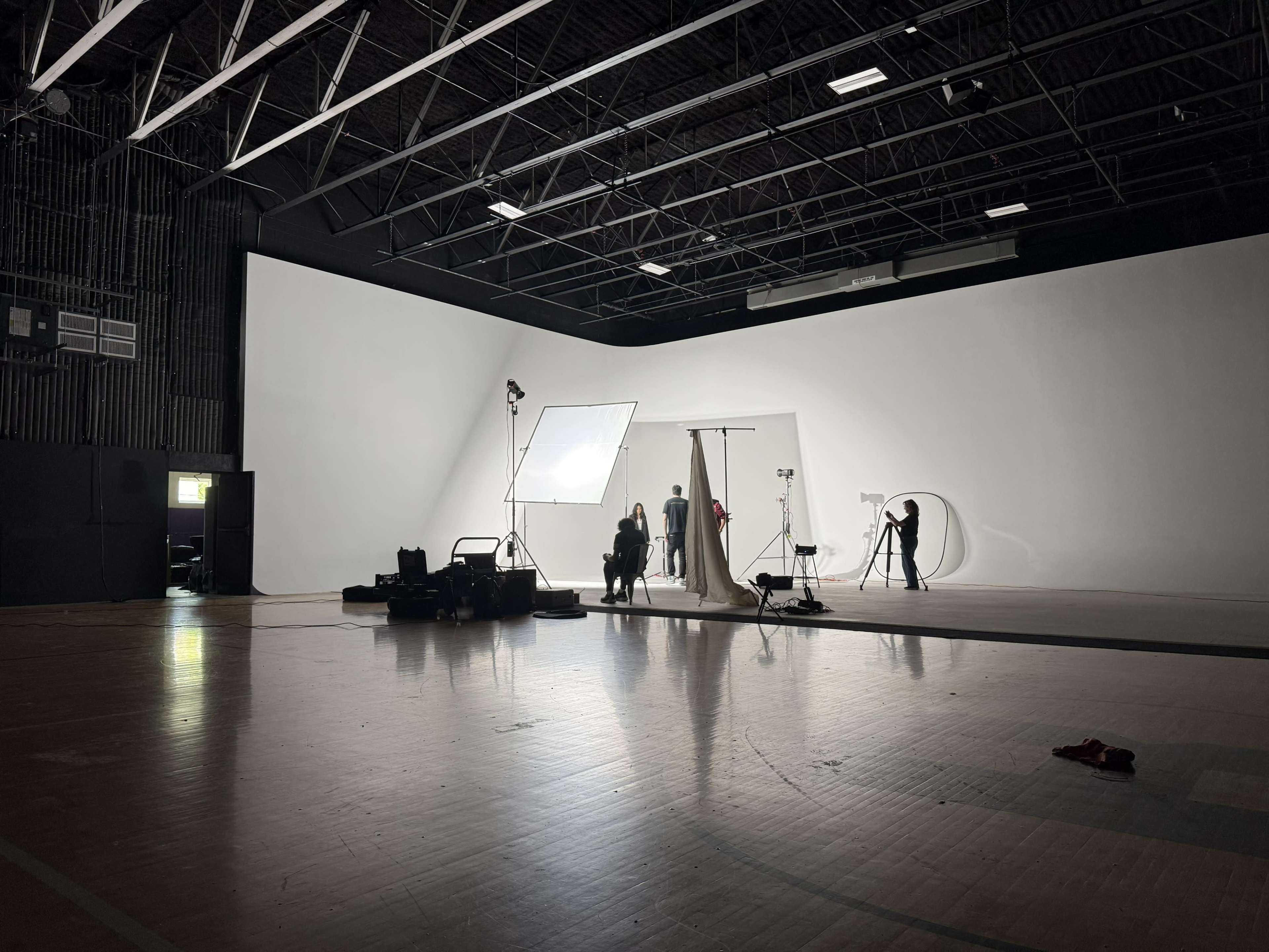 The image shows a spacious photography studio with a large white backdrop, equipment set up, and several individuals working on a photoshoot.