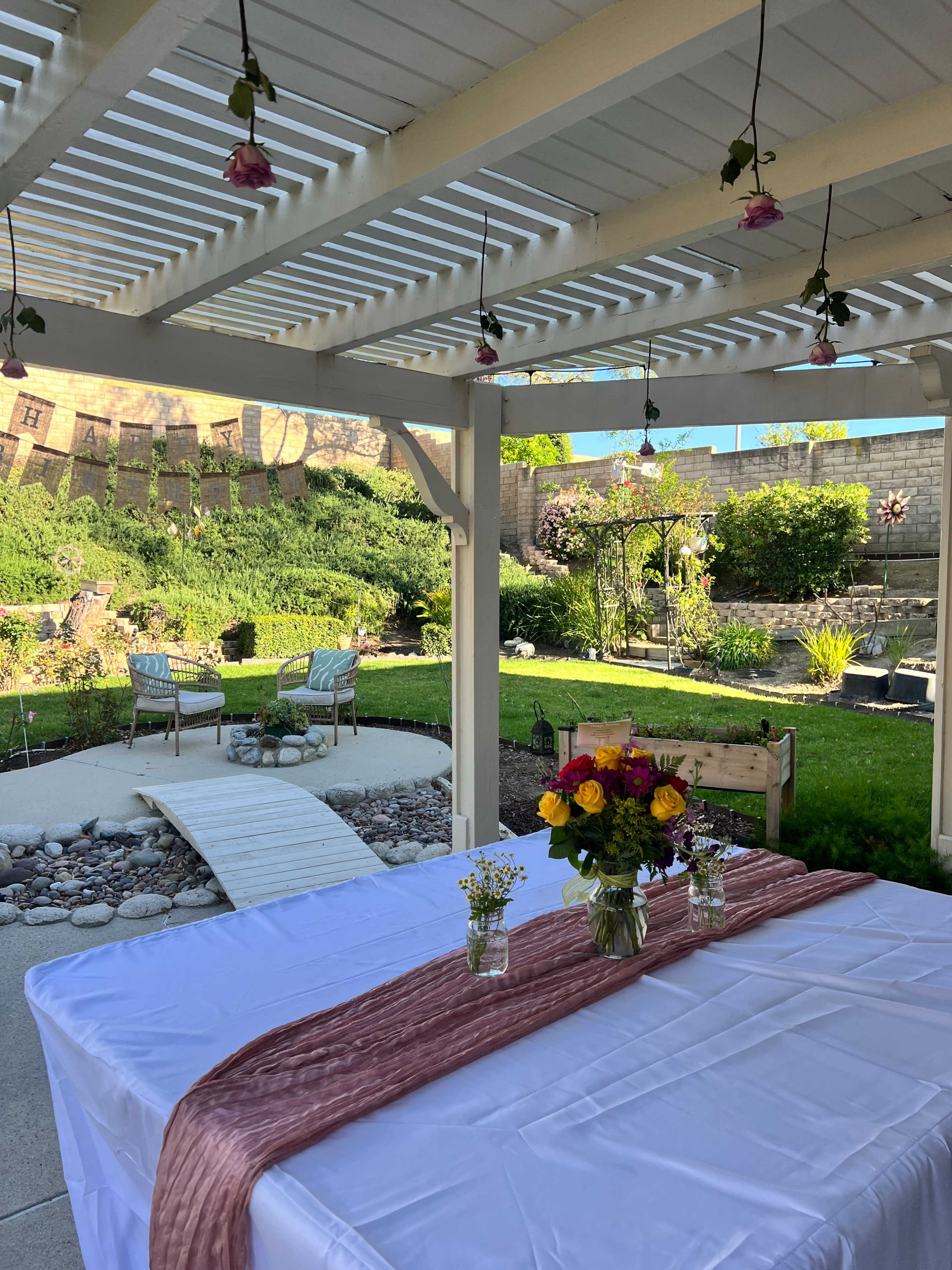 A neatly arranged outdoor patio features a table with a white cloth and a floral centerpiece, overlooking a landscaped garden with lounge chairs and a winding path.
