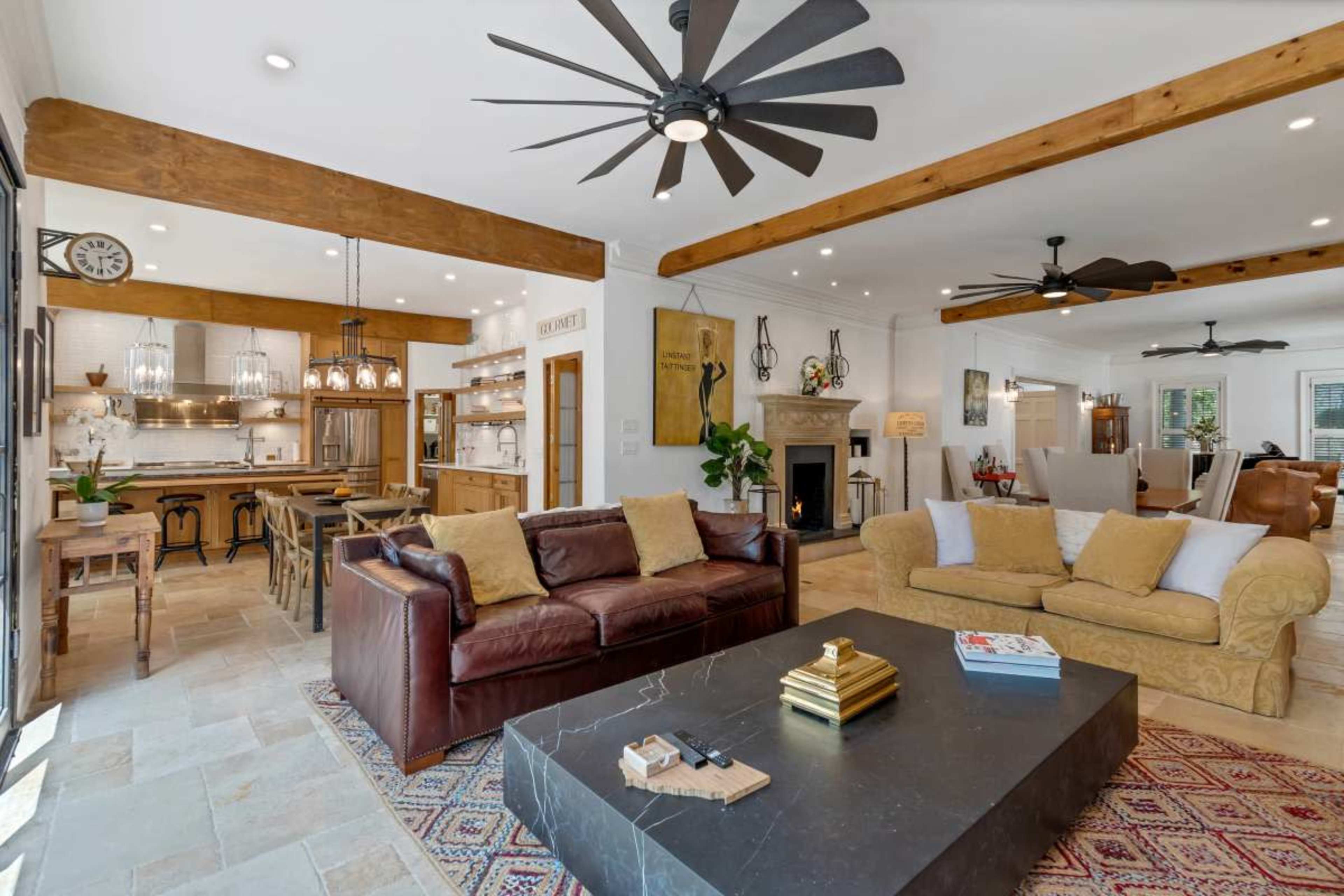 The image shows a modern living space with a brown leather sofa, a yellow couch, a coffee table, and an open kitchen area featuring wooden beams and stylish lighting fixtures.