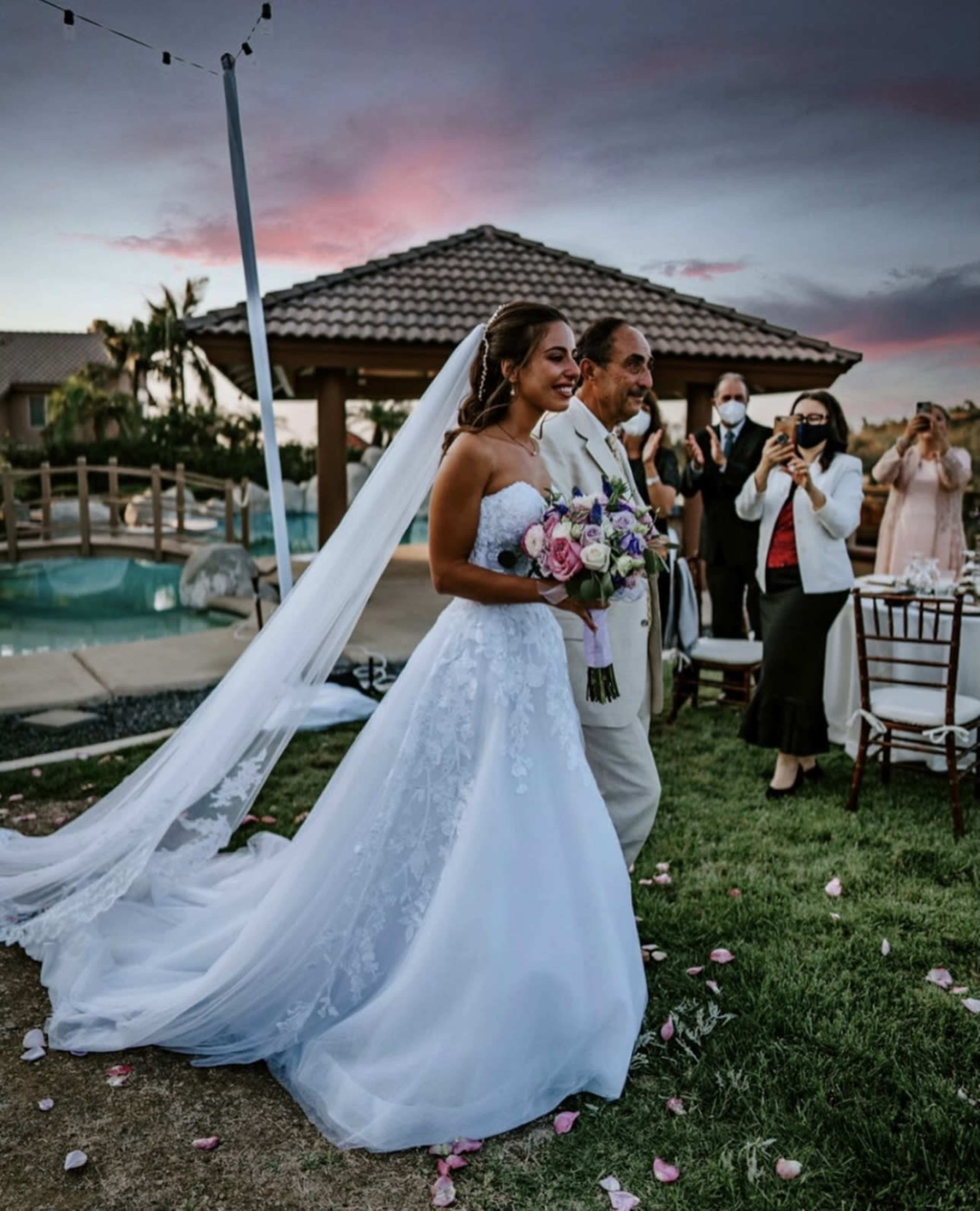 A bride in a white wedding dress holds a bouquet while walking with her father toward a gathering of guests during an outdoor ceremony at sunset.