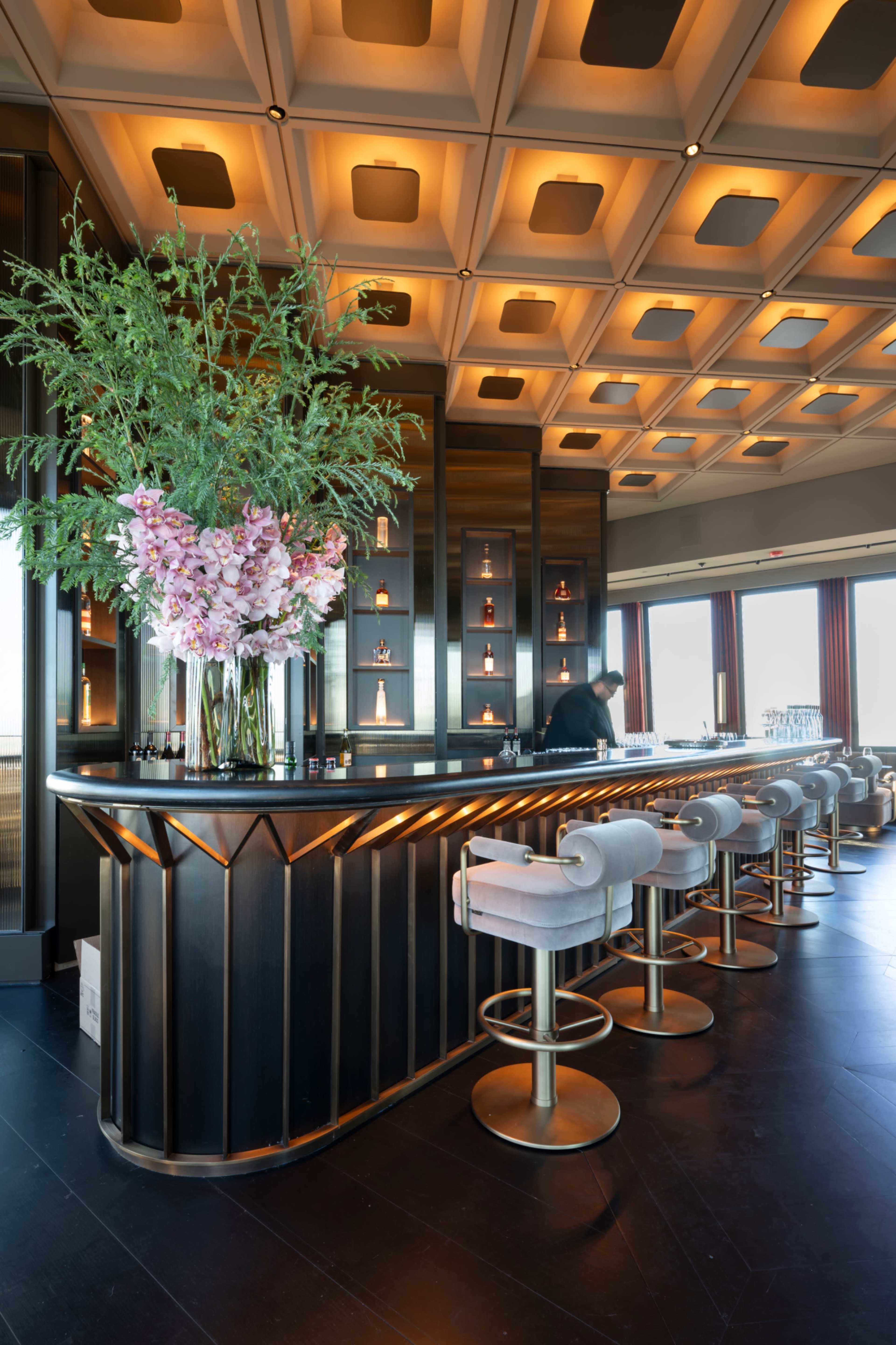 A sleek bar with a black and gold design is adorned with large floral arrangements and surrounded by white upholstered stools under a coffered ceiling.