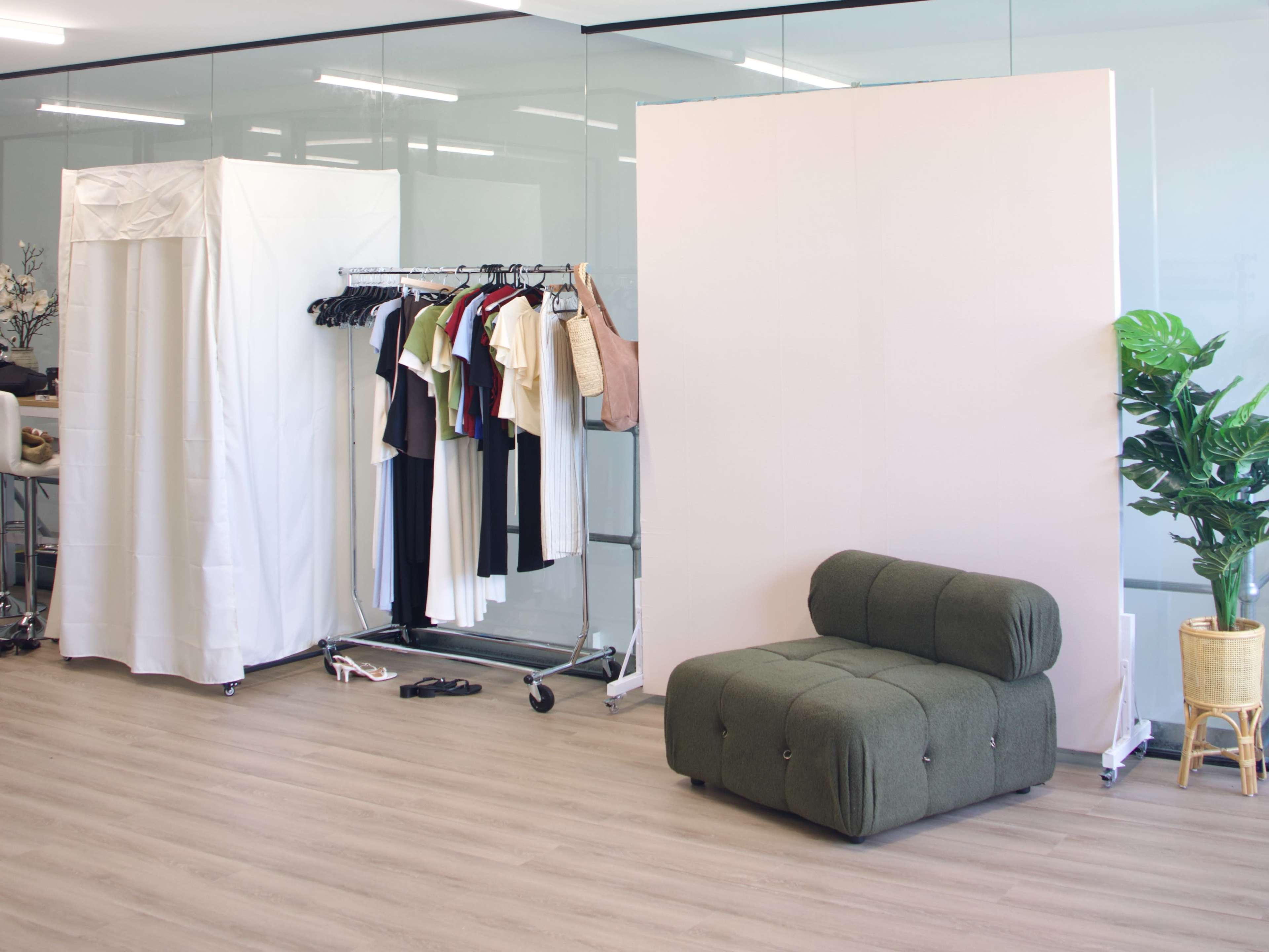 The image shows a modern studio space featuring a clothing rack with various garments, a portable changing area draped in white, a simple green couch, and a potted plant.