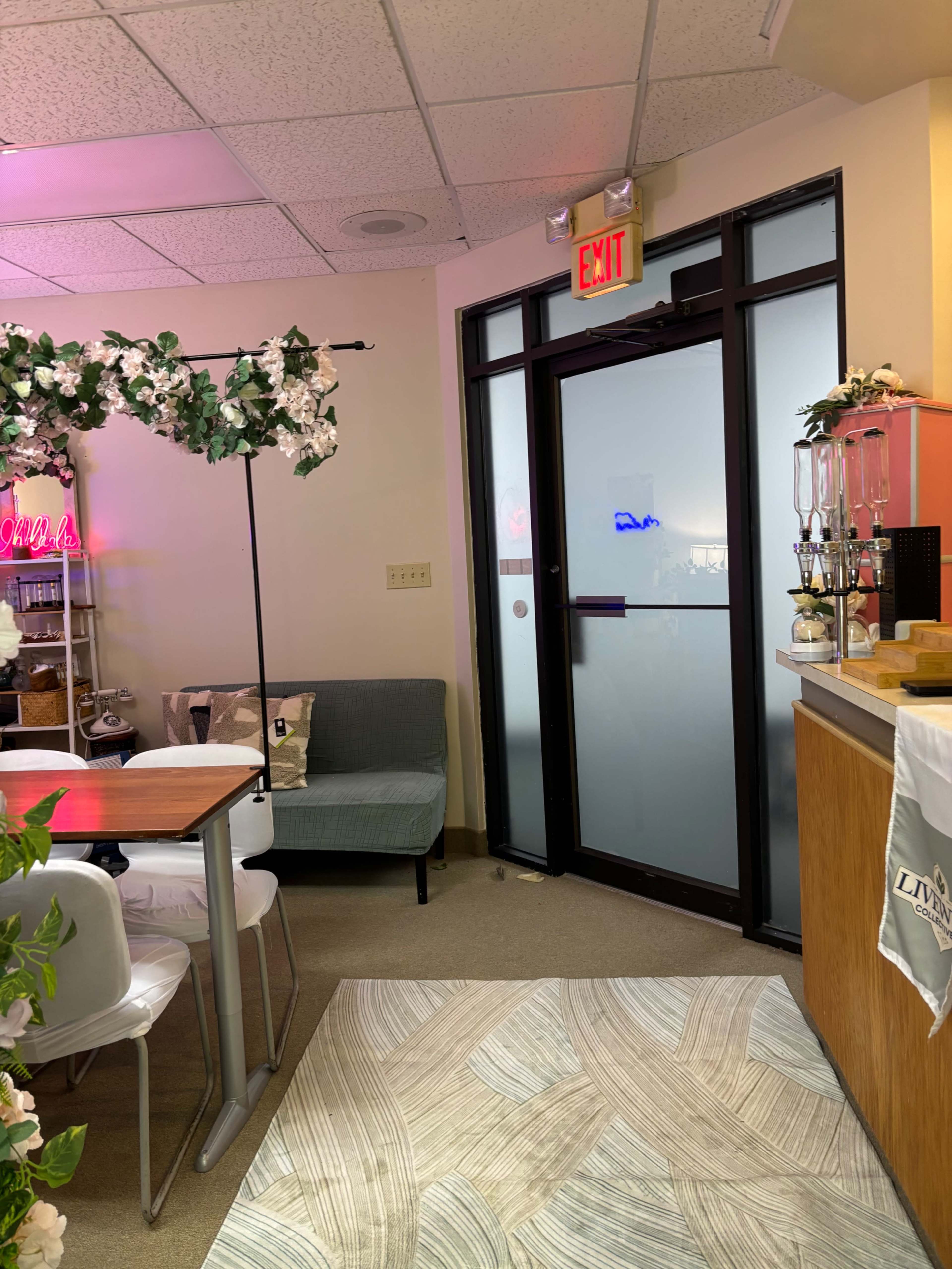 The image shows a cozy café interior with a counter, seating area, and a glass door leading outside, decorated with plants and neon signs.