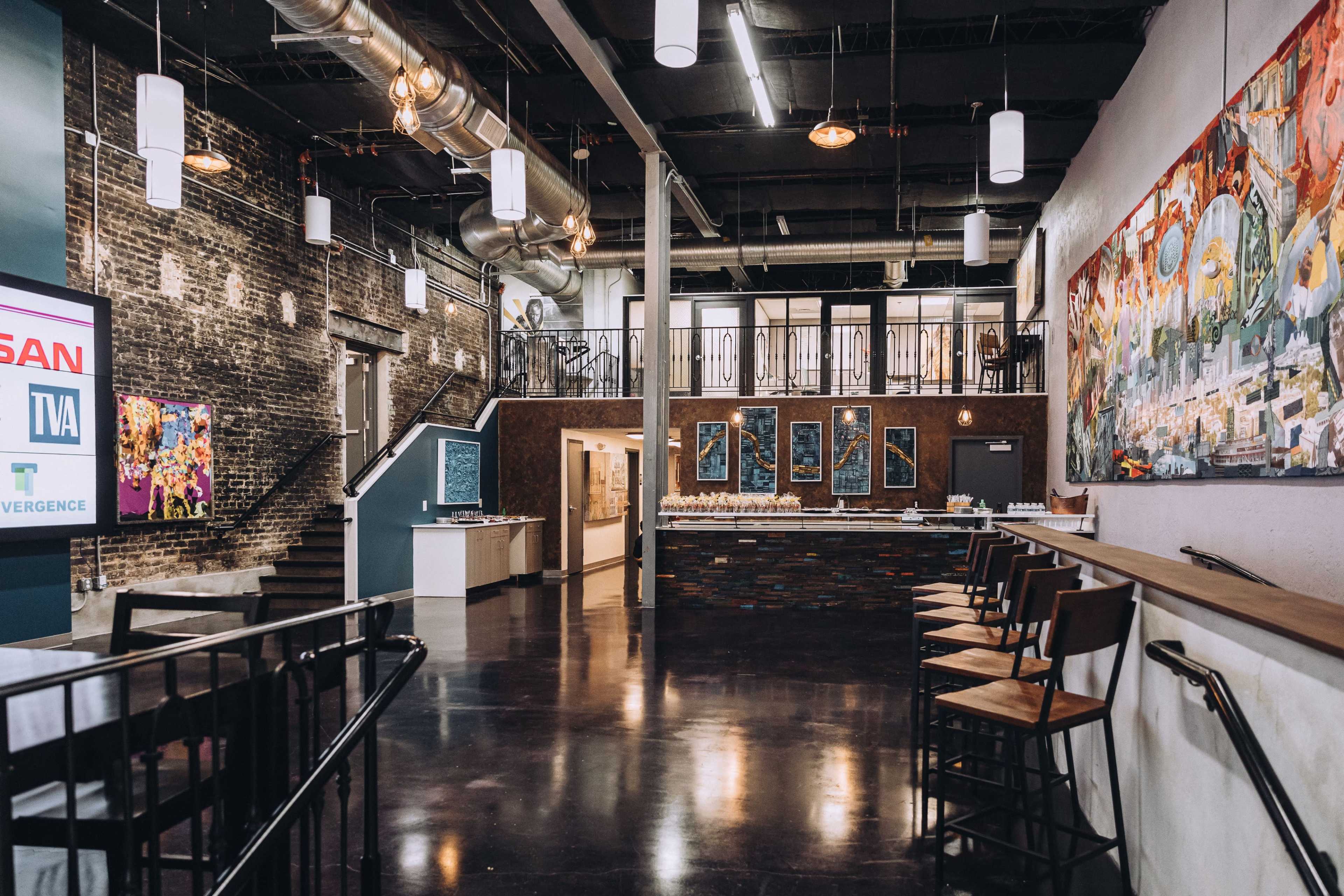 The image shows a modern, open interior of a restaurant or event space with exposed brick walls, high ceilings, and a mix of seating options along with artwork on the walls.
