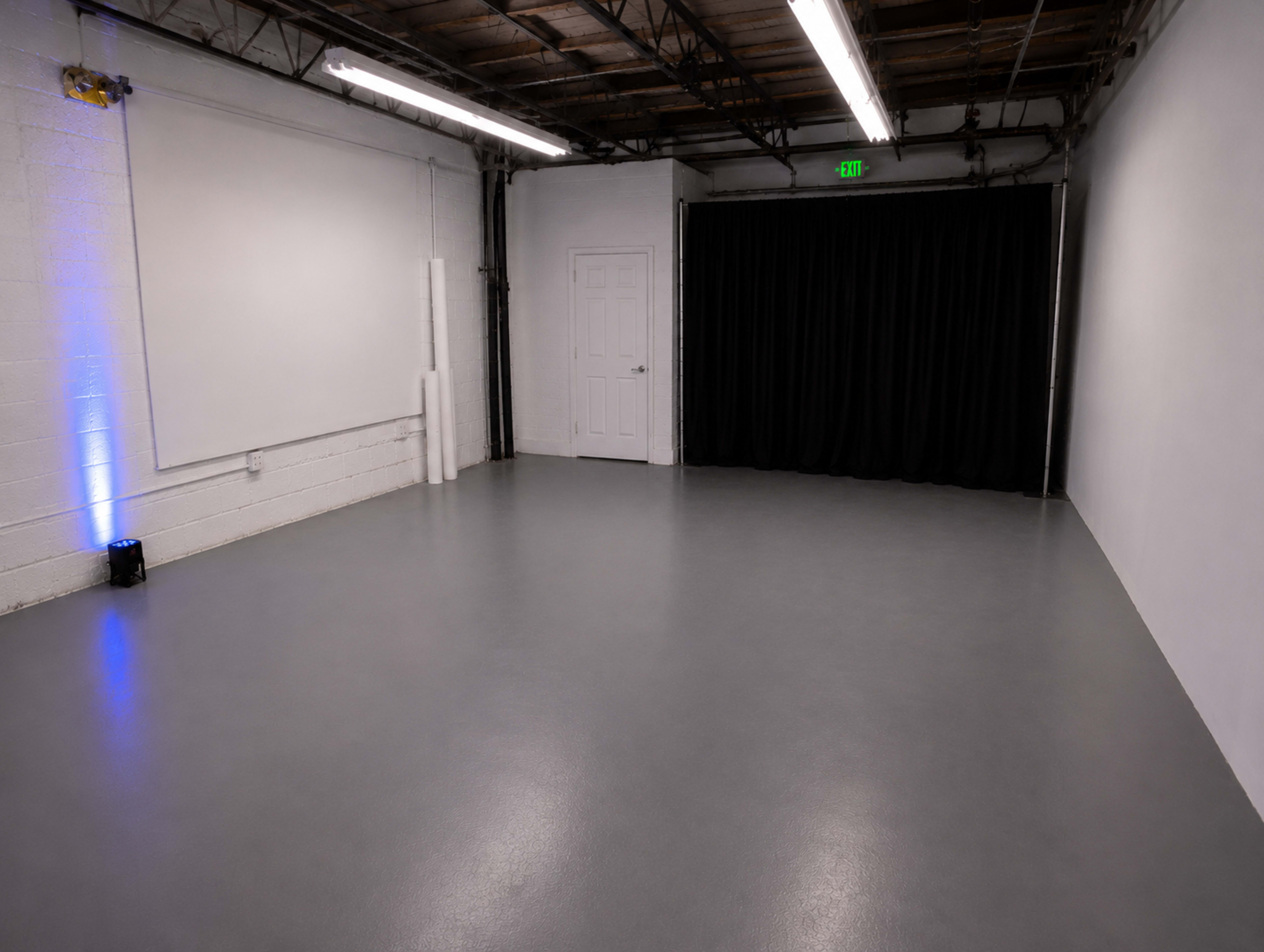 The image shows a spacious, empty room with gray flooring, white walls, and a black curtain on one side.