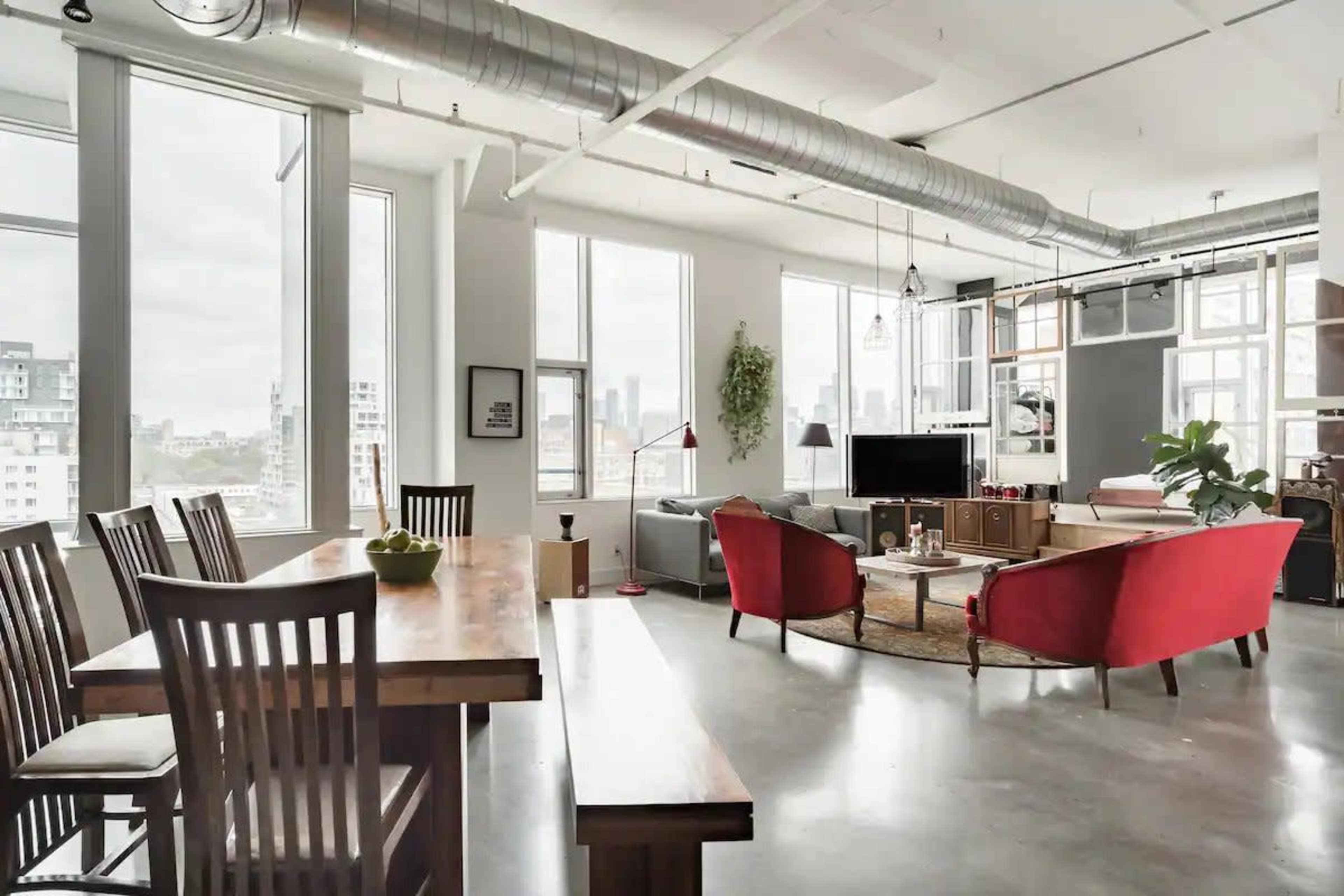 A modern loft-style living space with large windows, a dining table, and a seating area featuring red sofas.