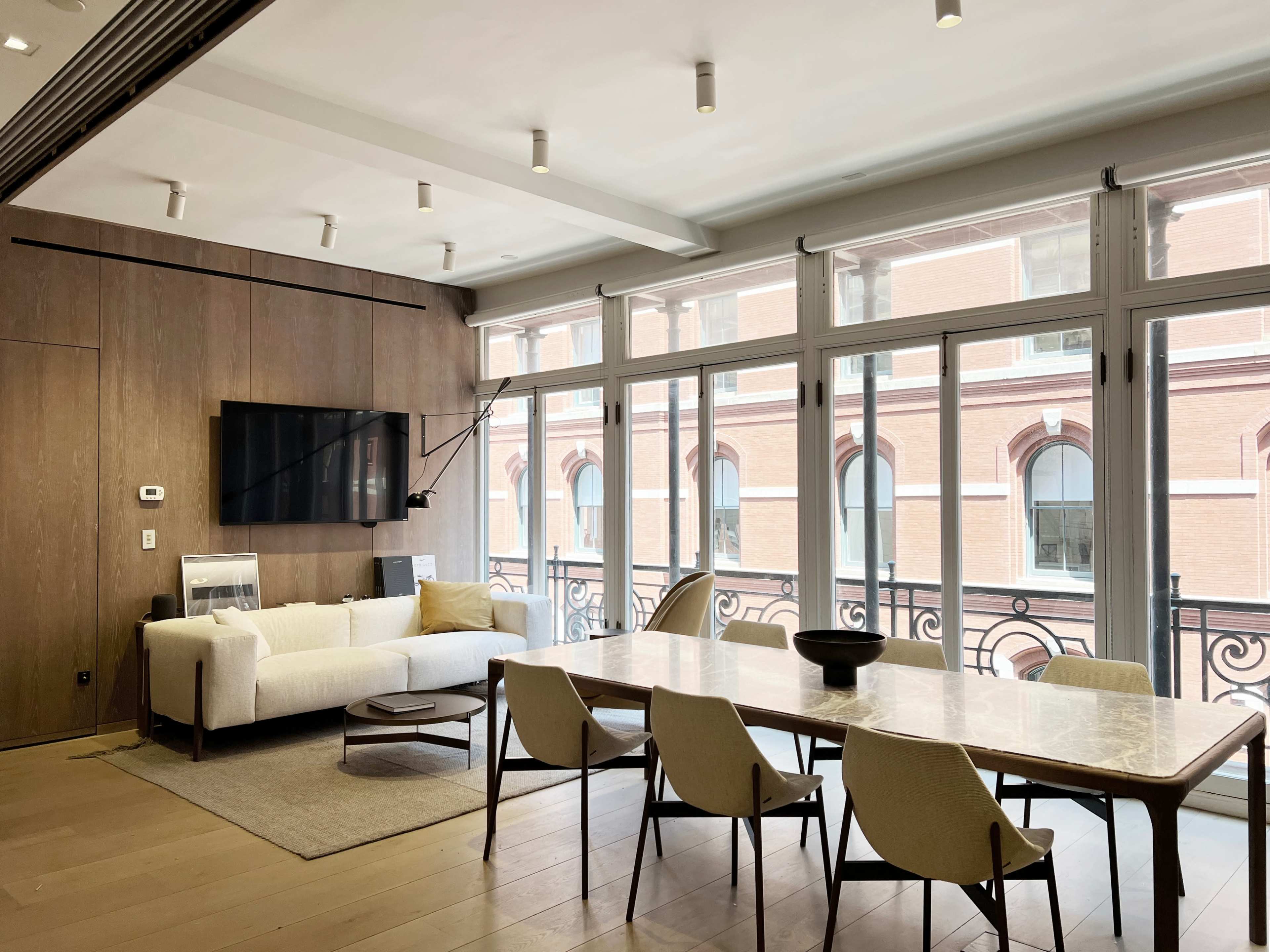 A modern living space with a white sofa, a wooden dining table surrounded by chairs, large windows, and a wall-mounted television.
