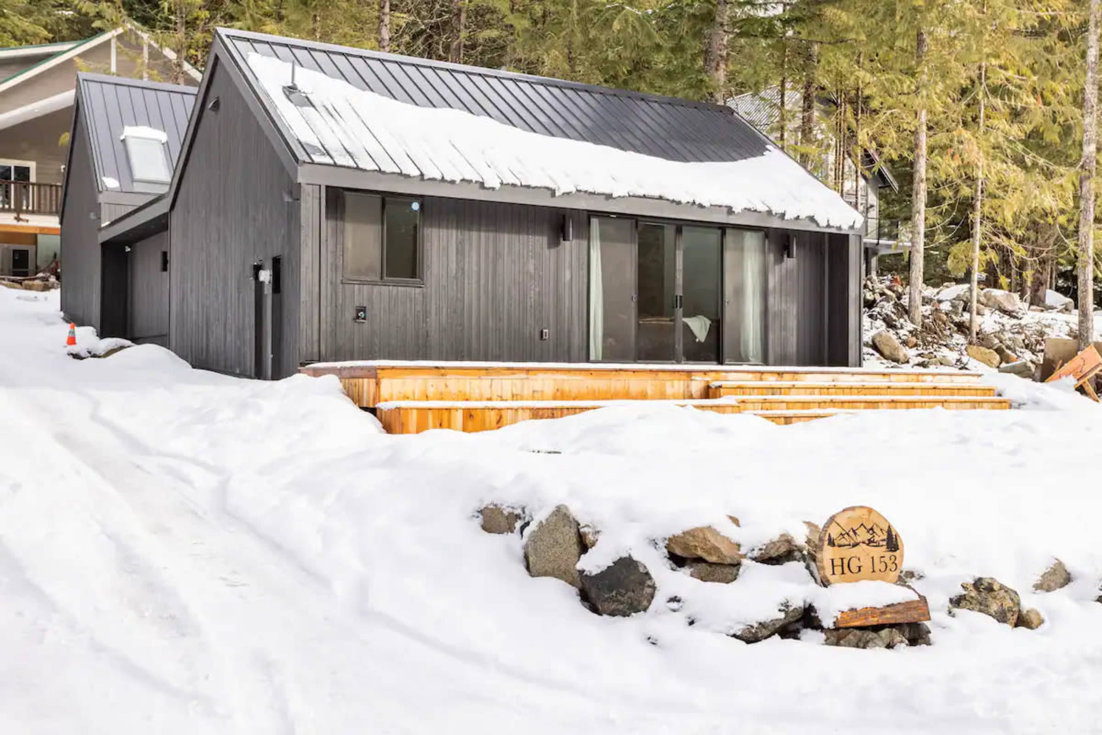 A modern black house with a metal roof sits on a snowy landscape, featuring wooden steps leading to the entrance.