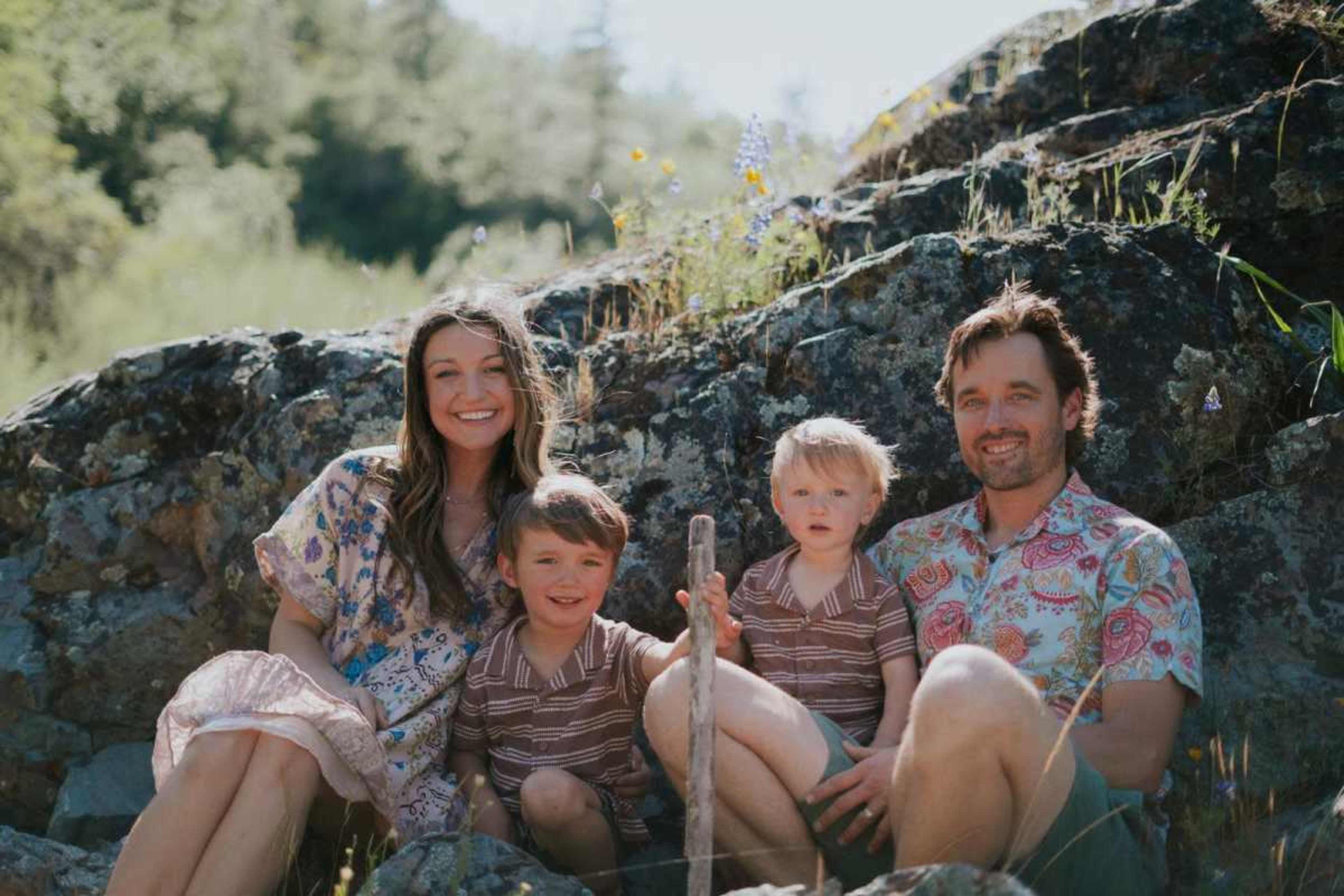 A family of four, including two young boys and their parents, sits on large rocks surrounded by greenery and wildflowers.