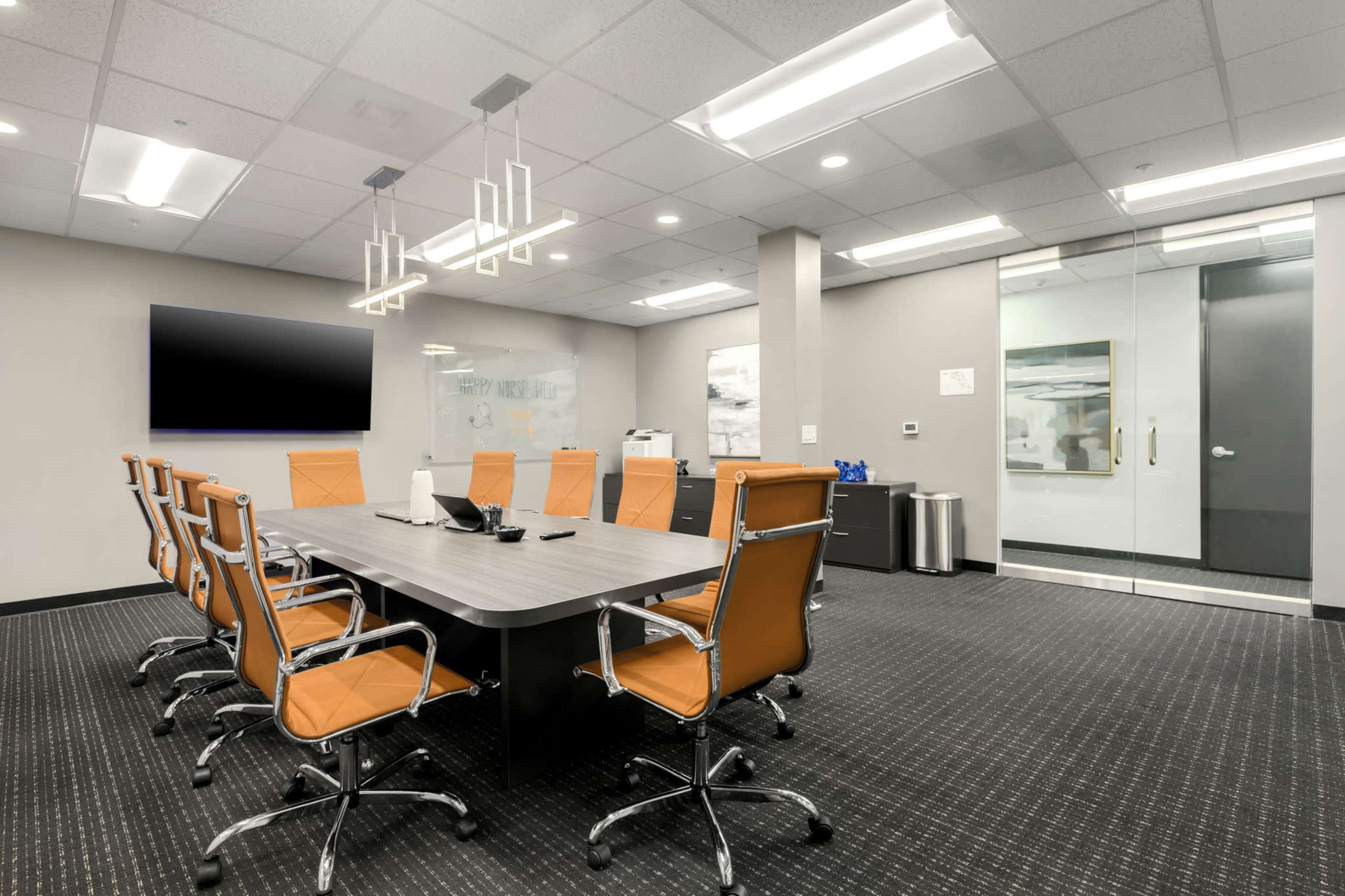 A modern conference room features a long table surrounded by orange chairs, with a large screen and glass doors leading to another area.