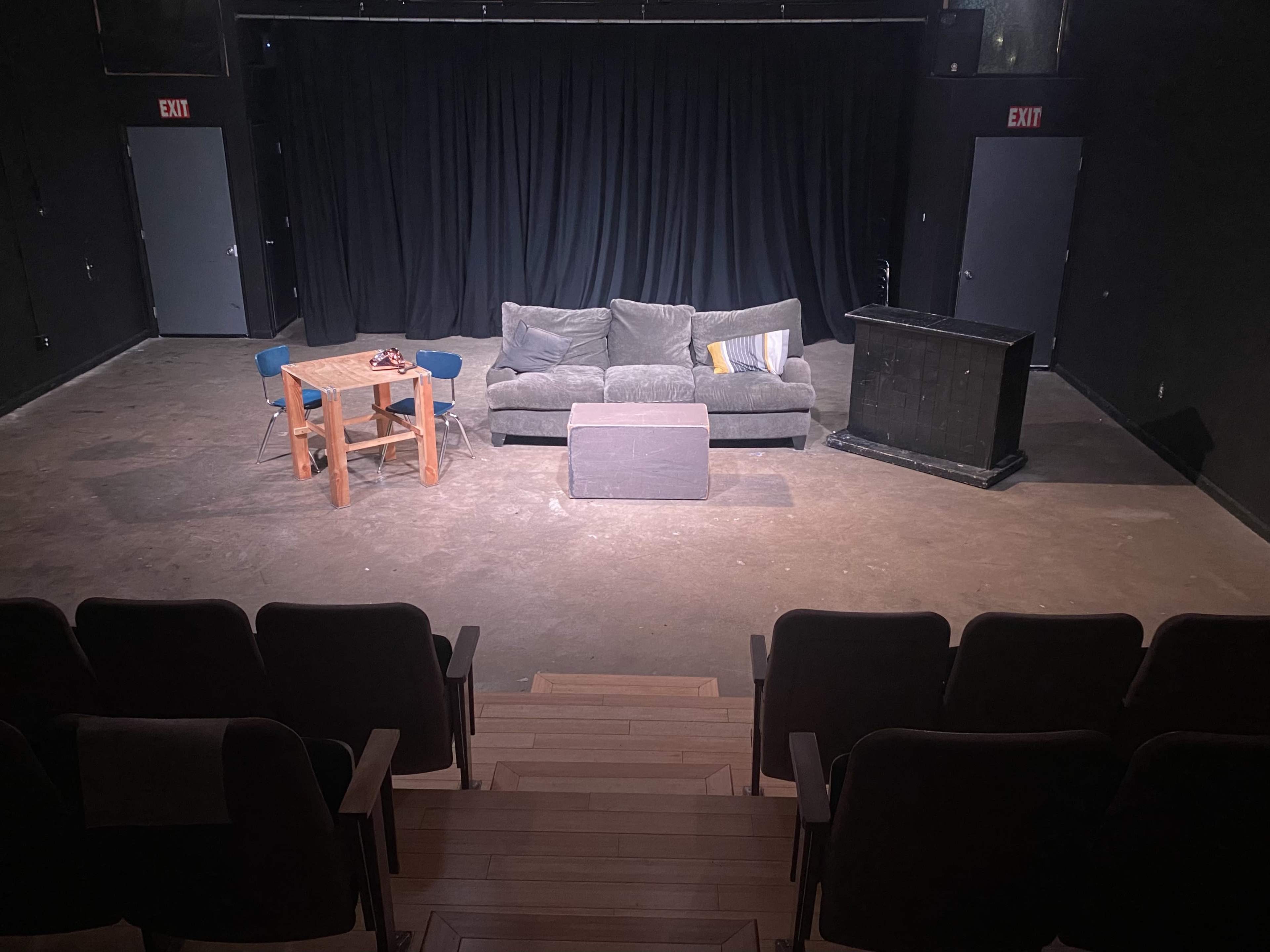 A small theater stage features a gray couch, a wooden table, a black podium, and a black curtain at the back.