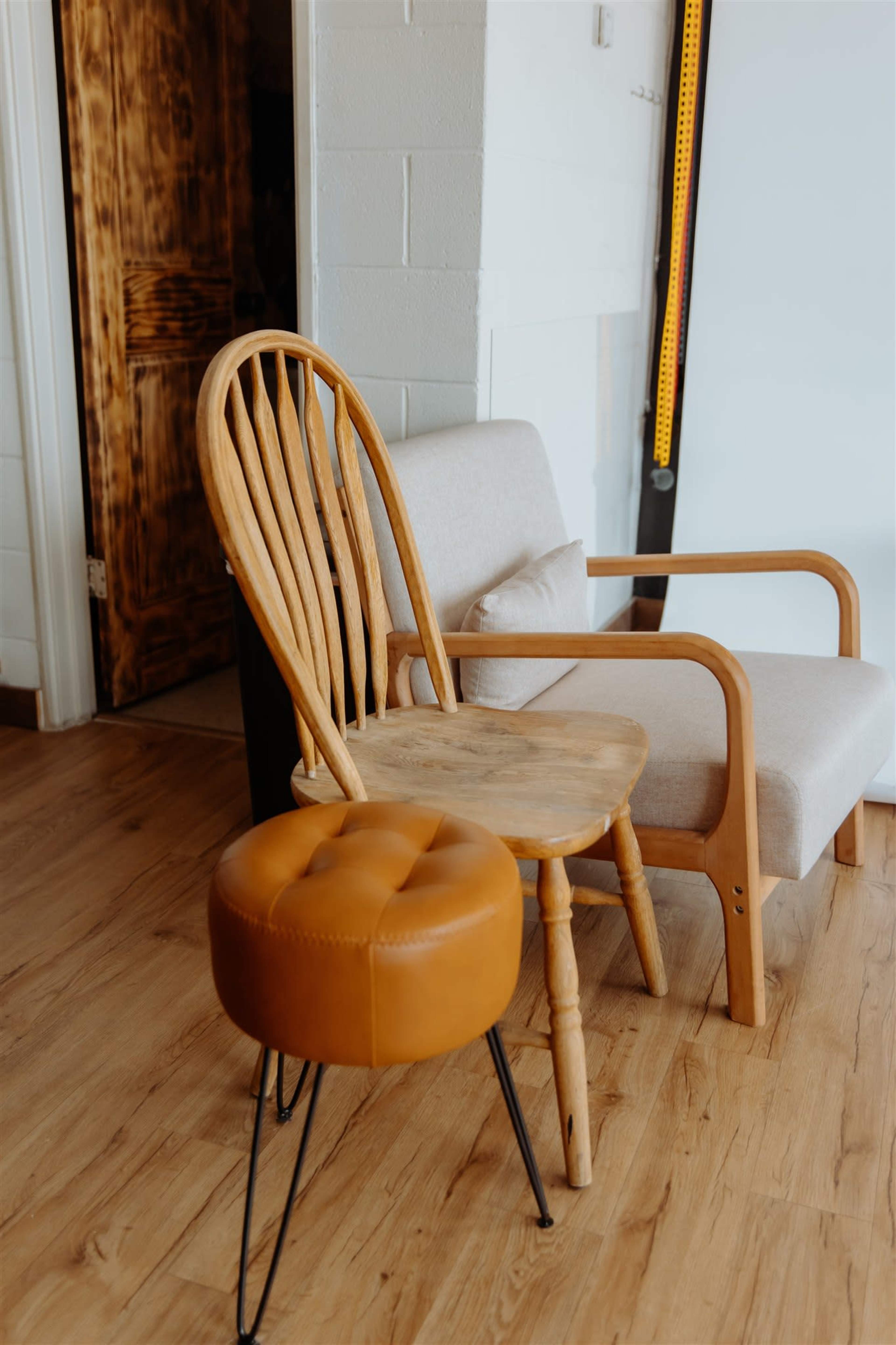 A wooden chair and a round leather ottoman are placed beside a light-colored sofa in a room with wooden flooring.