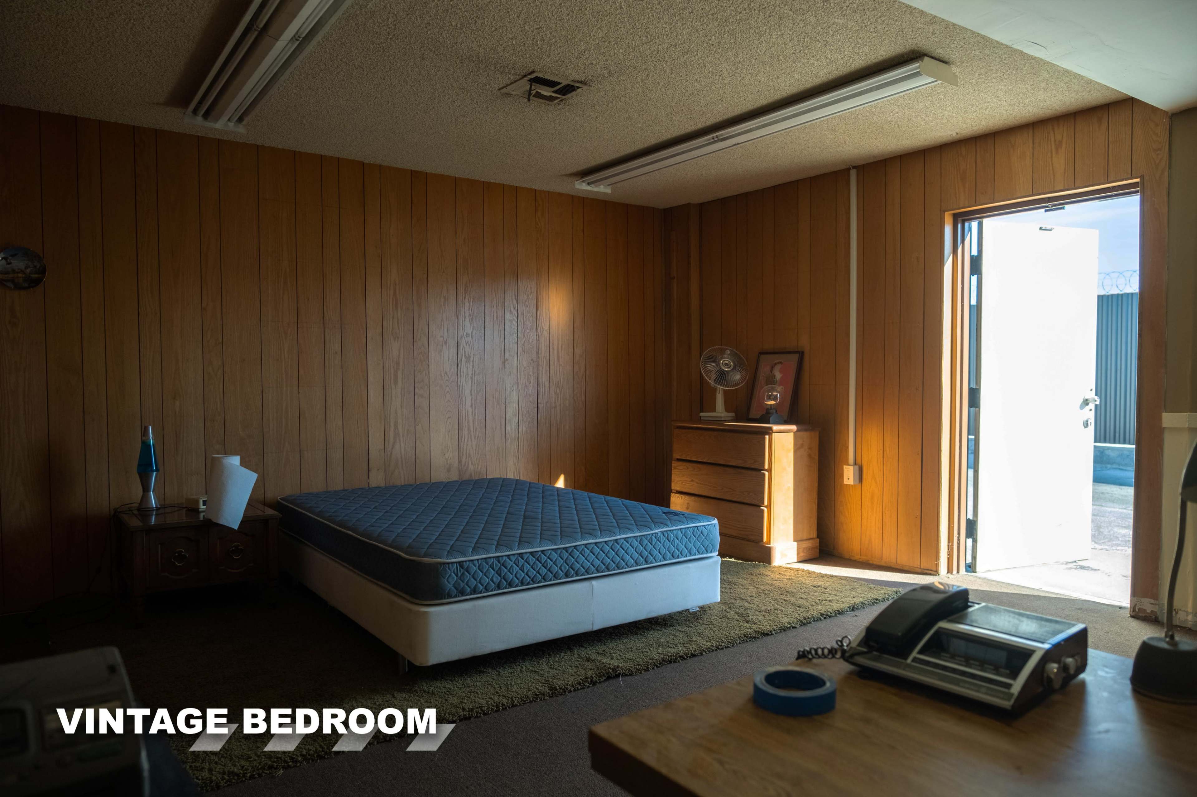 A vintage bedroom features a bare mattress on a wooden floor, a wooden dresser, and an open door revealing an external area.