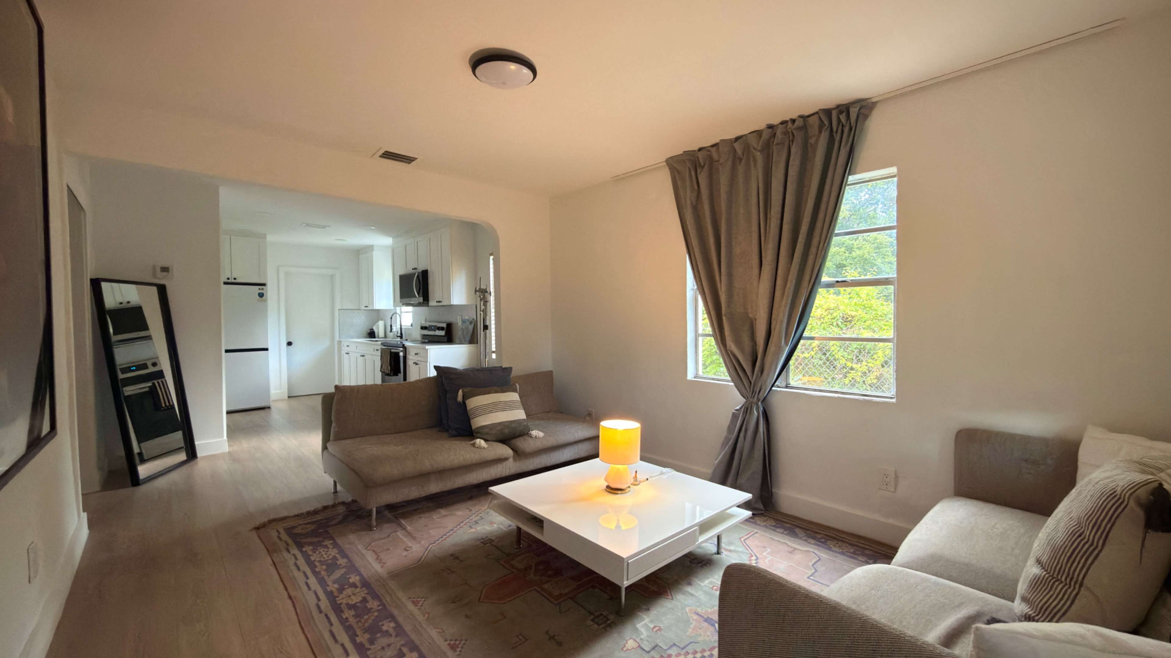 The image shows a modern living room with a couch, a coffee table, and large windows allowing natural light, connecting to a kitchen area in the background.