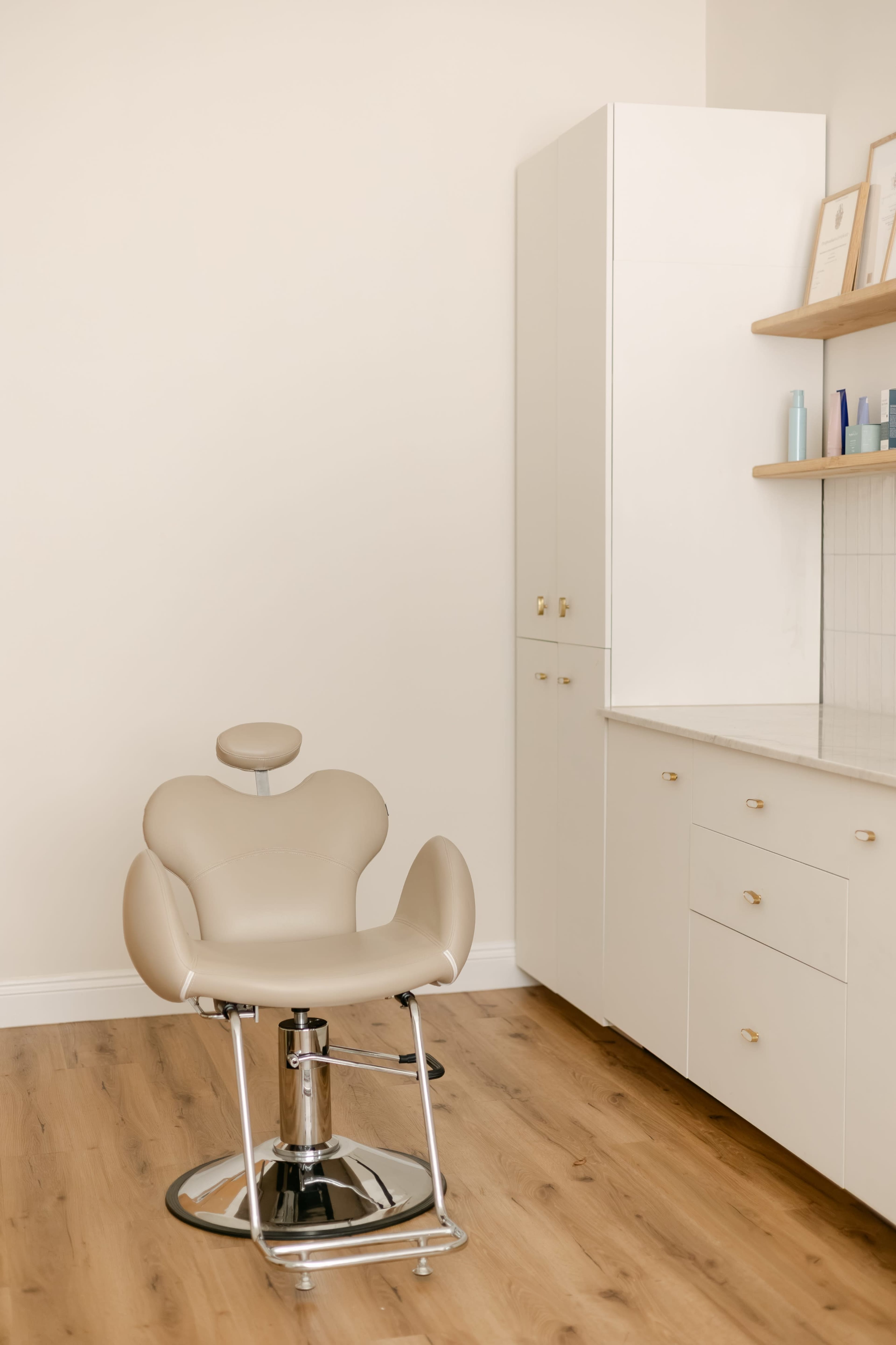 The image shows a empty salon chair positioned in a minimalistic room with light wood flooring and white cabinetry.