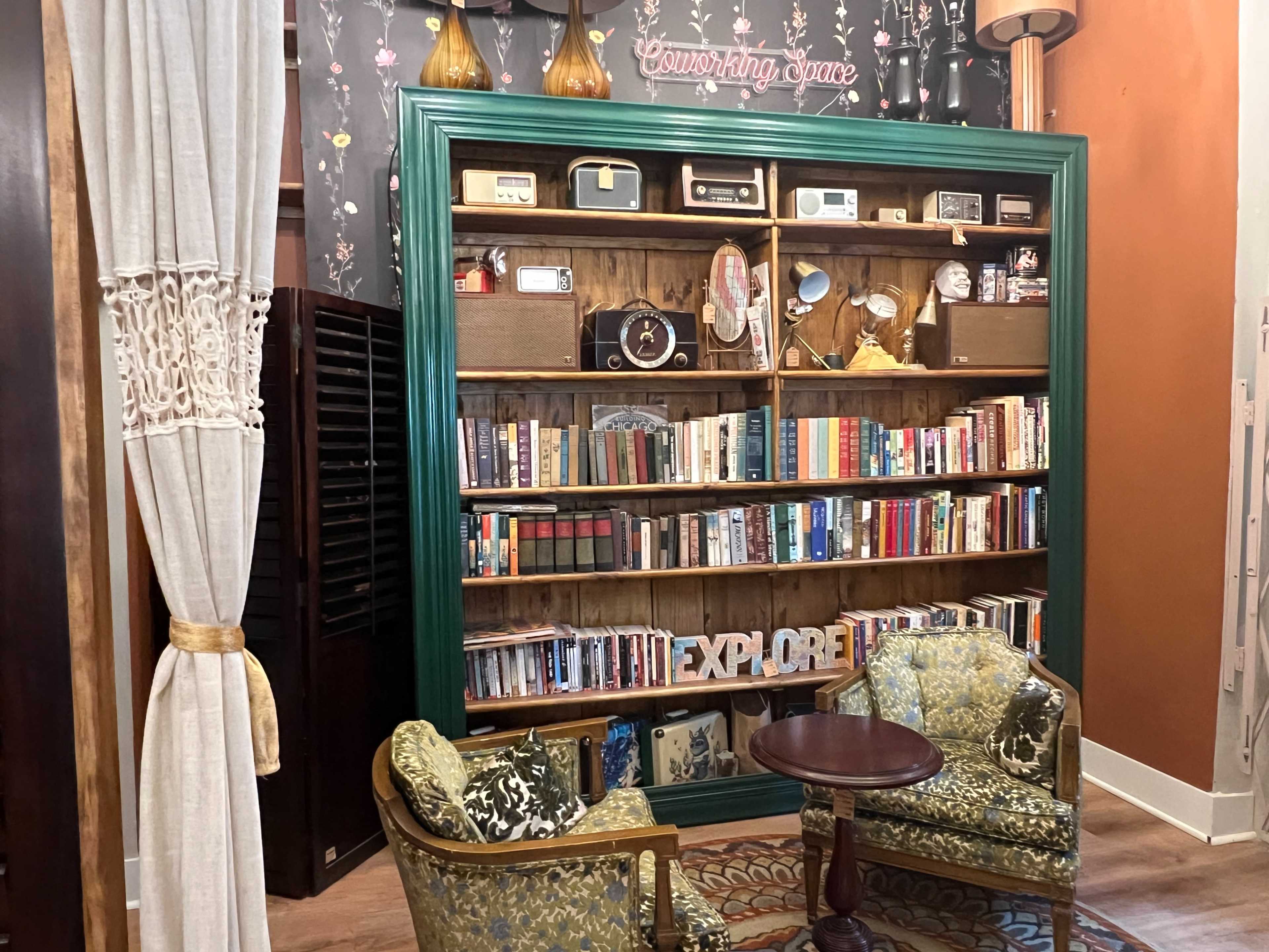 A wooden bookshelf filled with books and vintage items is flanked by two patterned armchairs and a small table in a cozy reading nook.