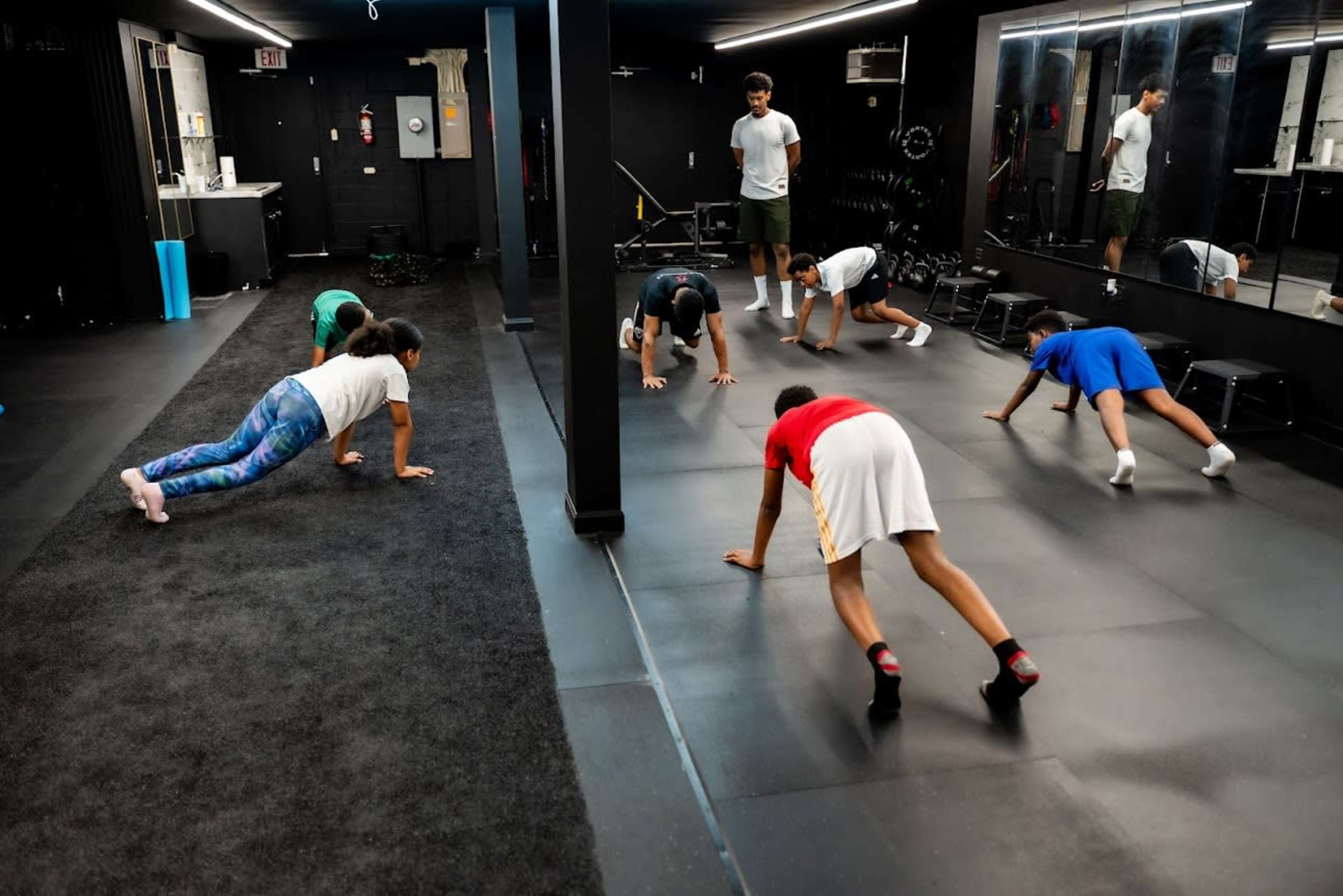 A group of children performs exercise drills in a fitness studio with black walls and carpeting.
