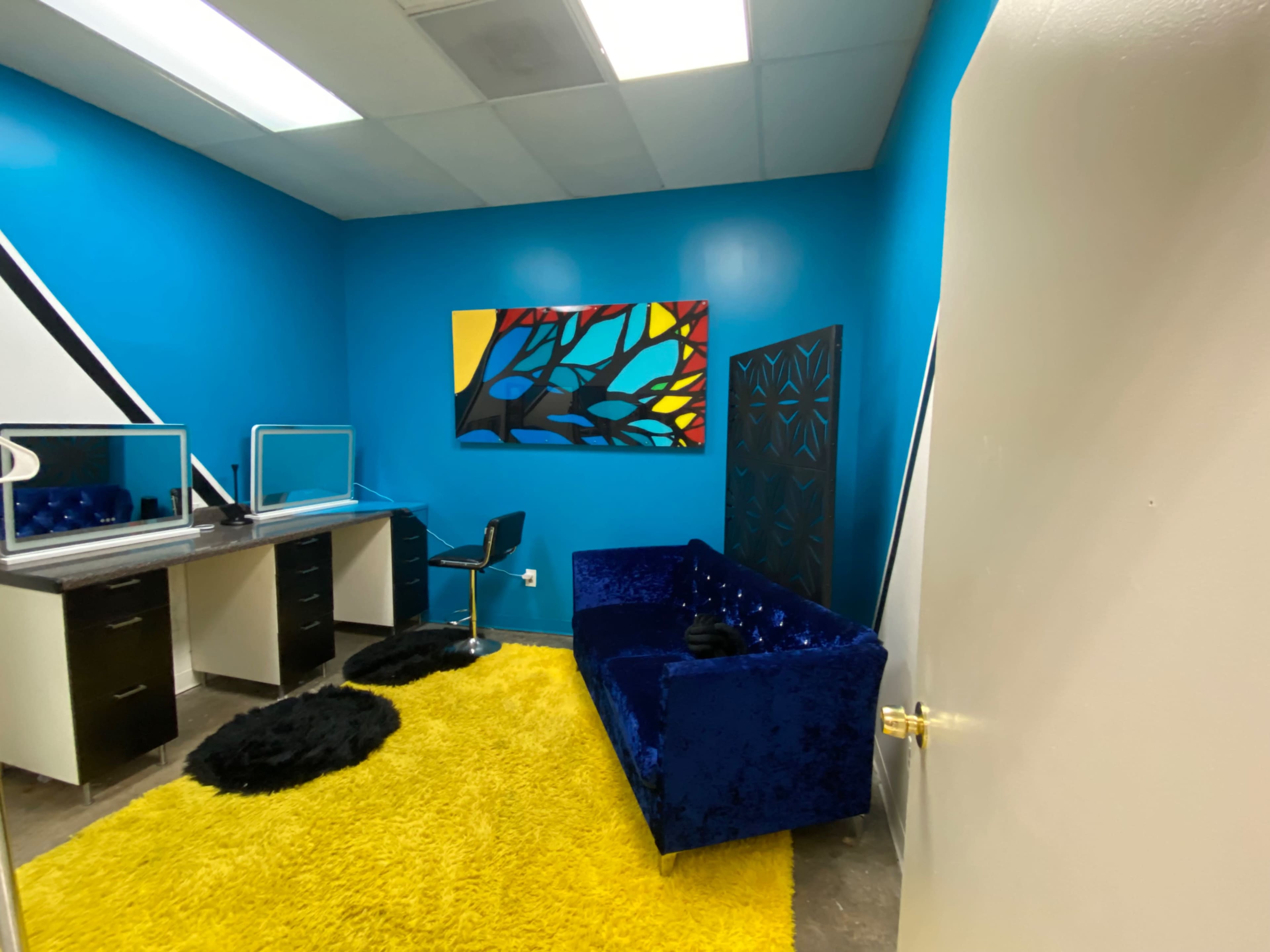 The image shows a small office space featuring a blue wall, a black and gold abstract painting, a blue velvet couch, and a desk with a mirror.