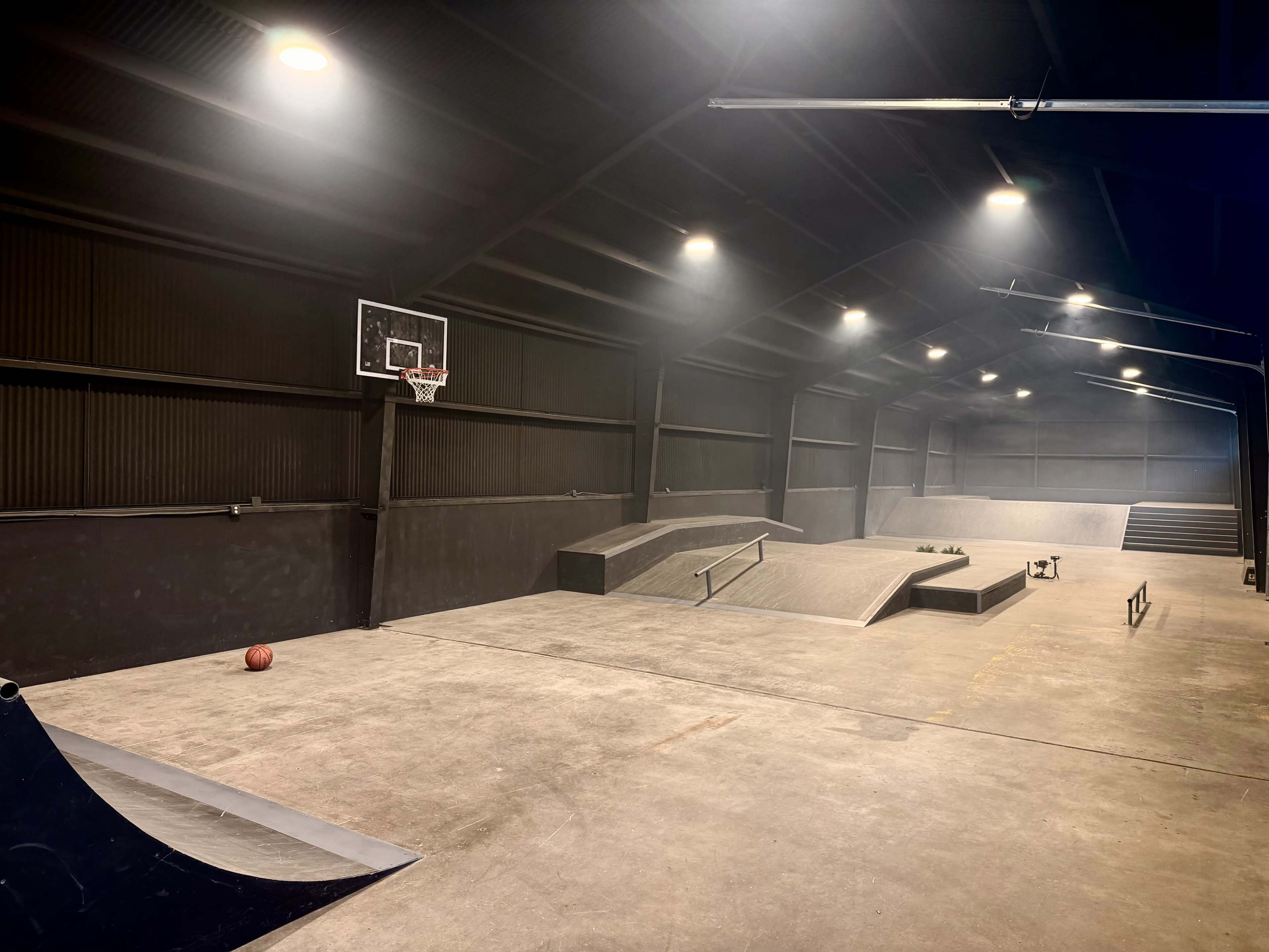 The image shows an indoor skate park featuring ramps, a basketball hoop, and scattered equipment under dim lighting.