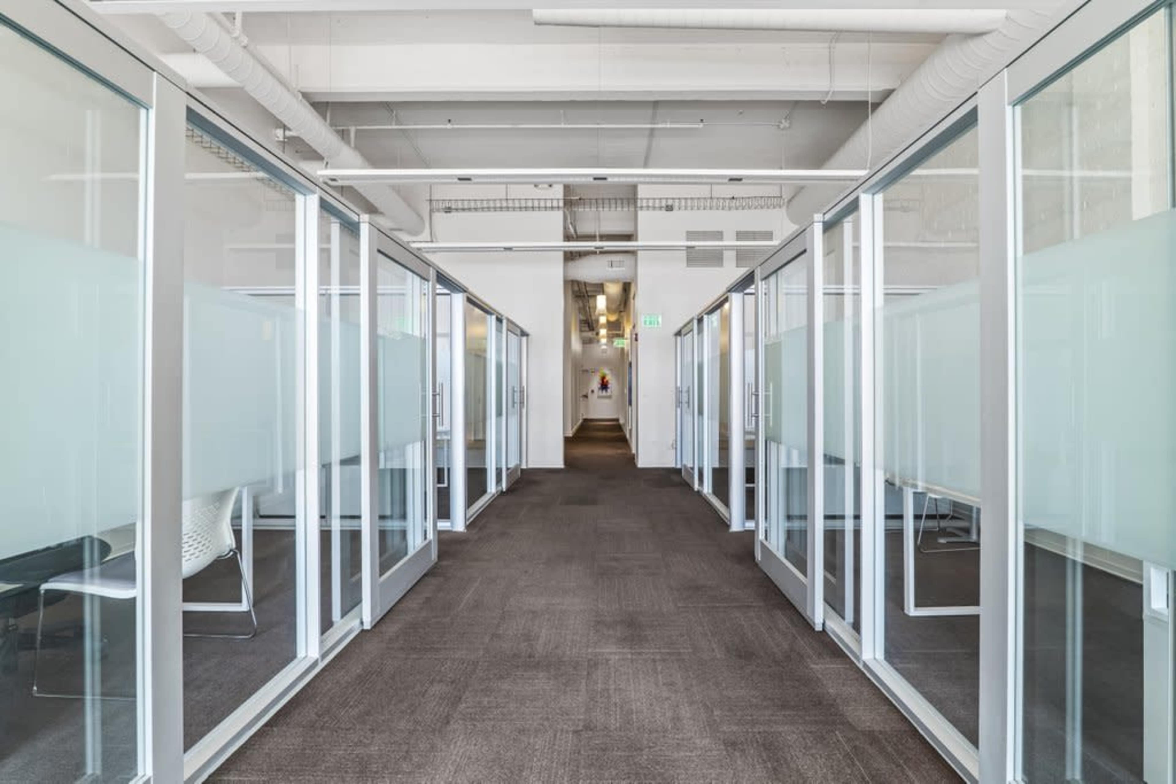 The image shows a modern office corridor lined with glass-walled meeting rooms on either side.