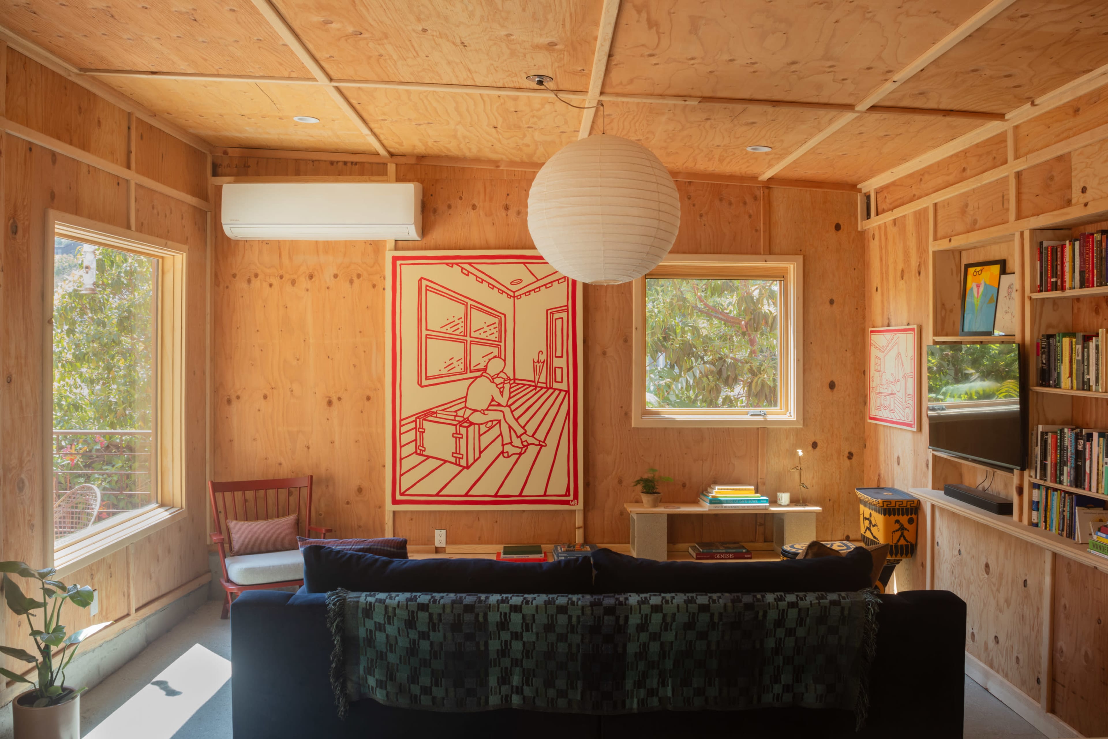The image depicts a modern, wooden interior living room featuring a large artwork, natural light from windows, and a black sofa.