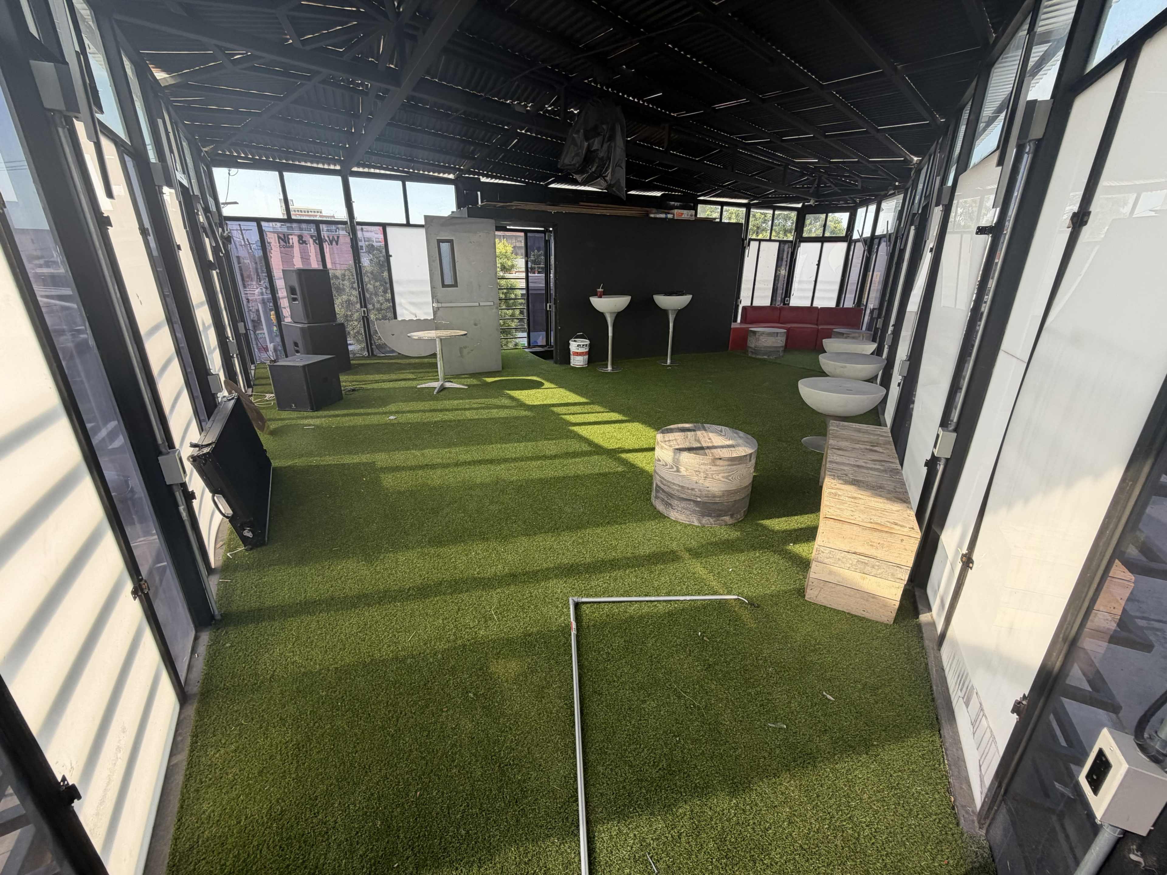 The image shows an empty rooftop space with artificial grass flooring, modern furniture, and large windows allowing natural light.