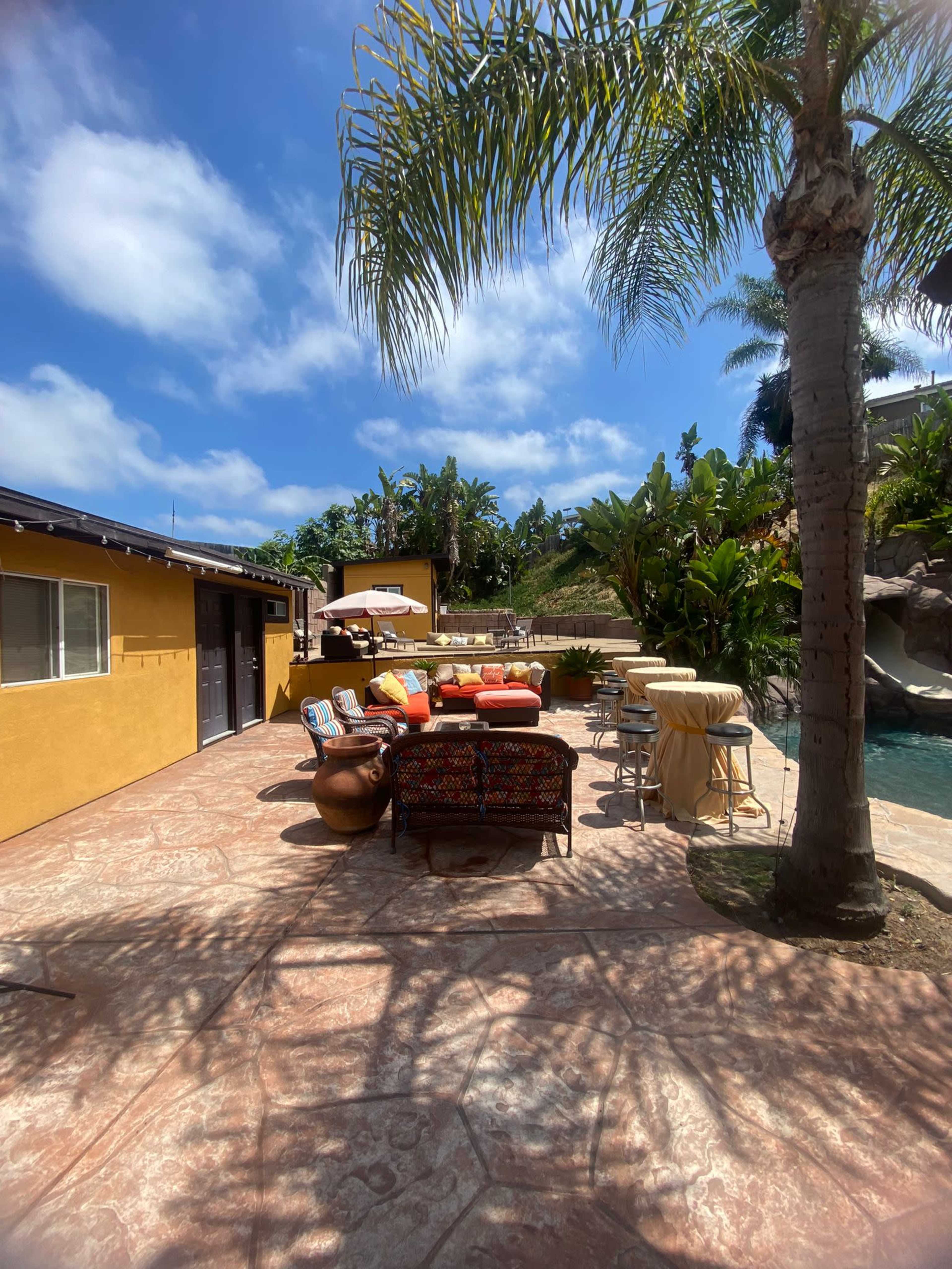 A patio area with colorful seating arrangements and a pool surrounded by tropical plants and trees under a partly cloudy sky.