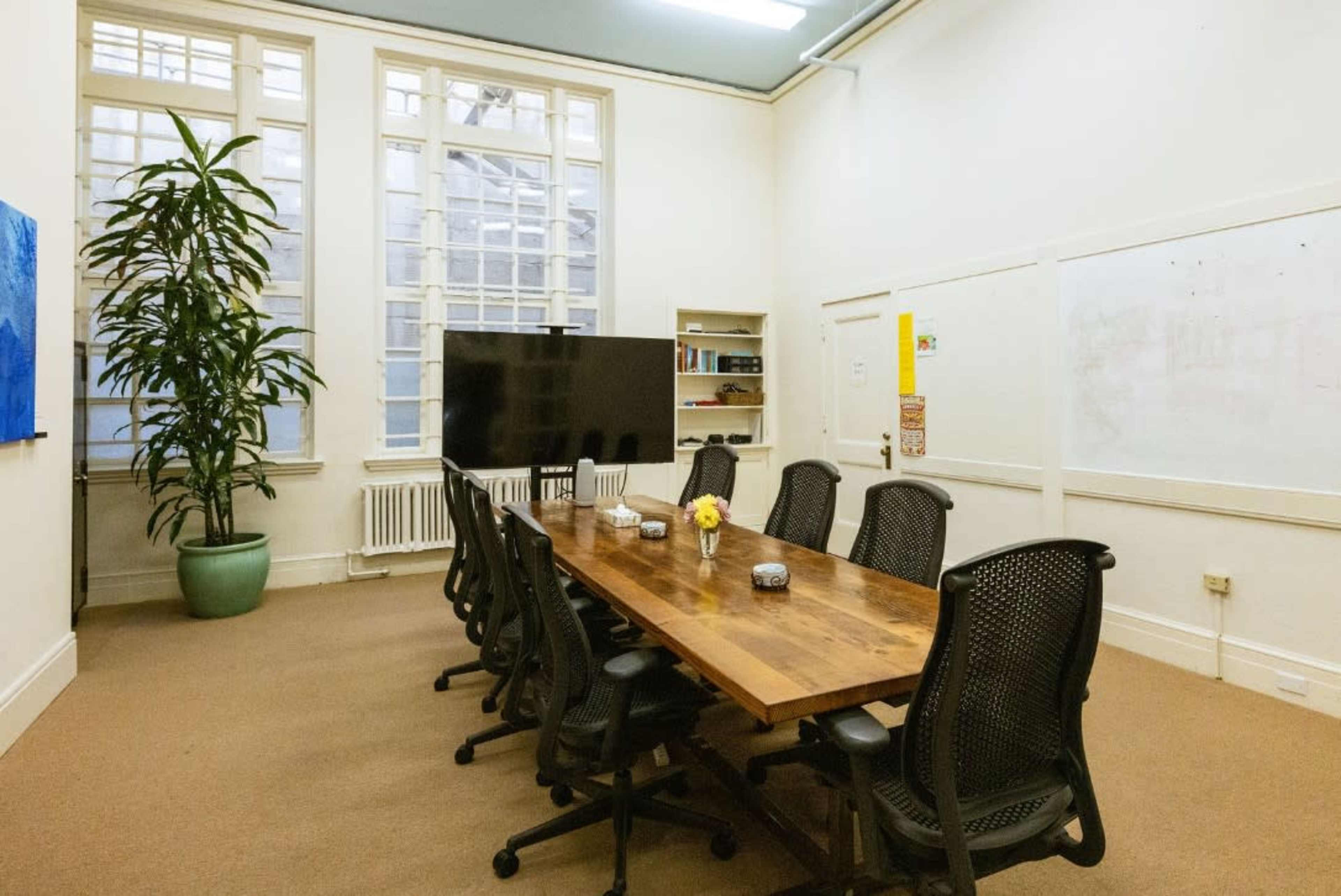 A conference room with a large wooden table surrounded by black ergonomic chairs, a potted plant in the corner, and a large screen against one wall.