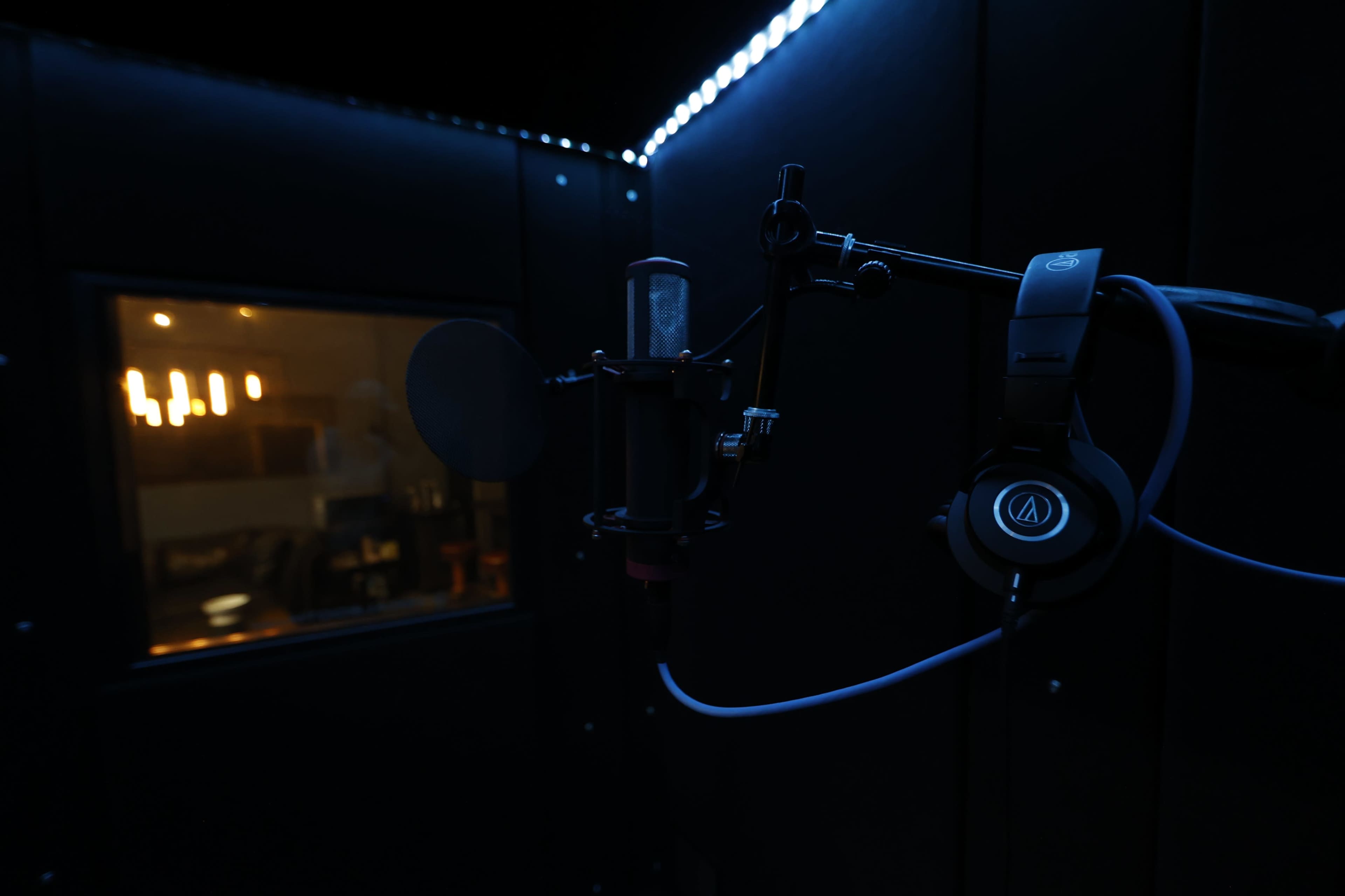 A microphone is positioned on a stand with headphones nearby inside a dark sound booth.