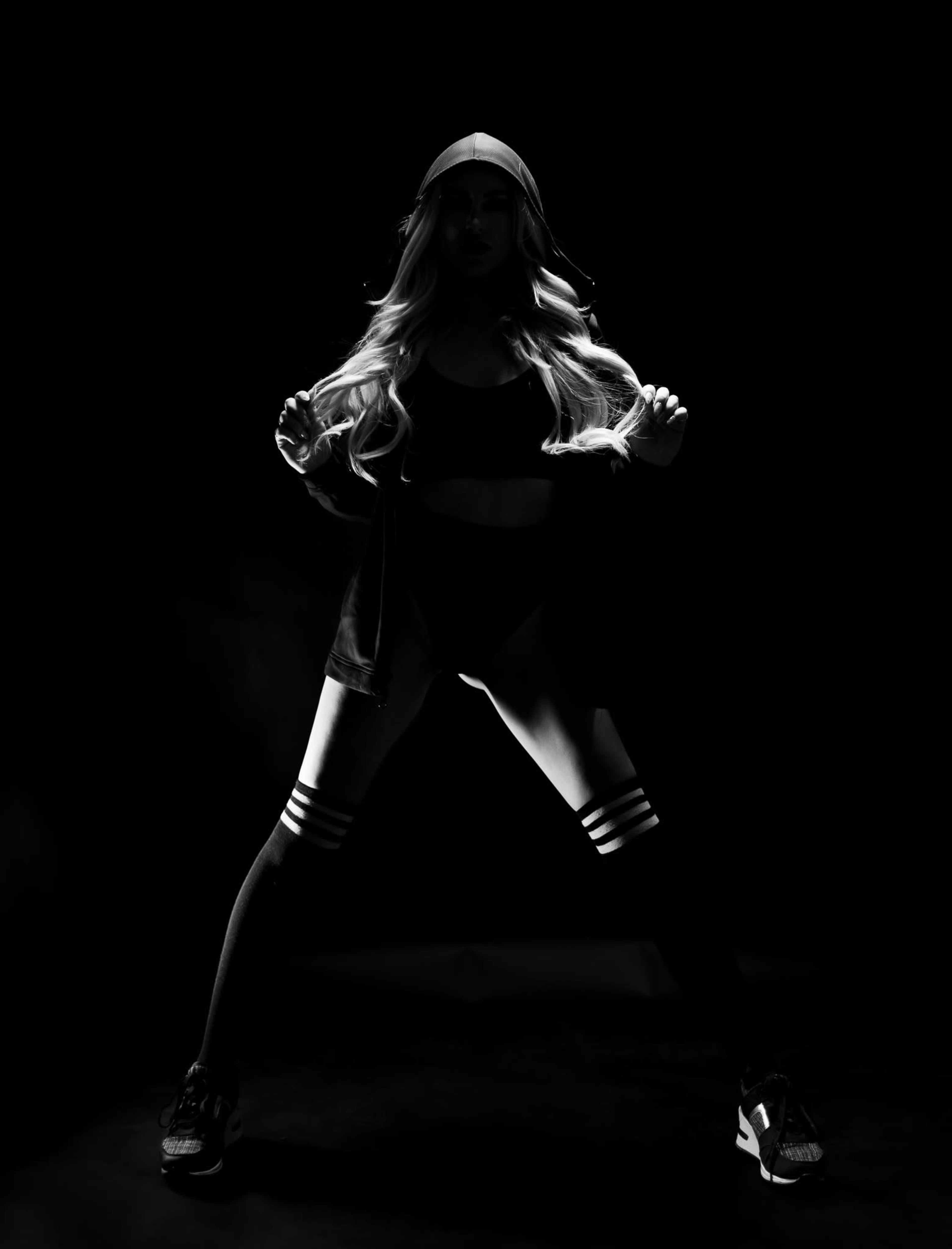 A person with long hair stands in a dark setting, wearing athletic clothing and striking a pose.
