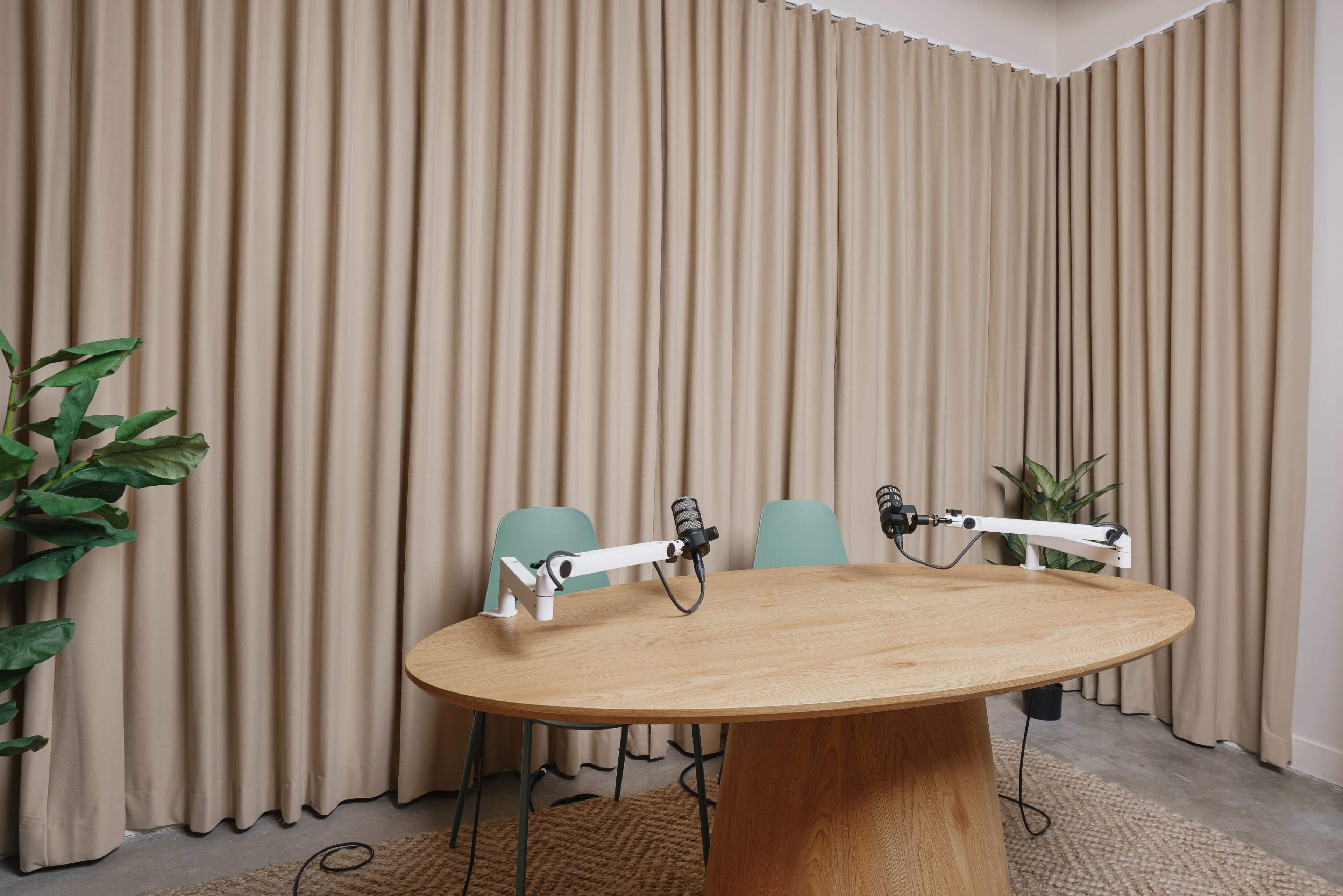 A round wooden table with two chairs and microphone stands is set up in a room with beige curtains and a potted plant.
