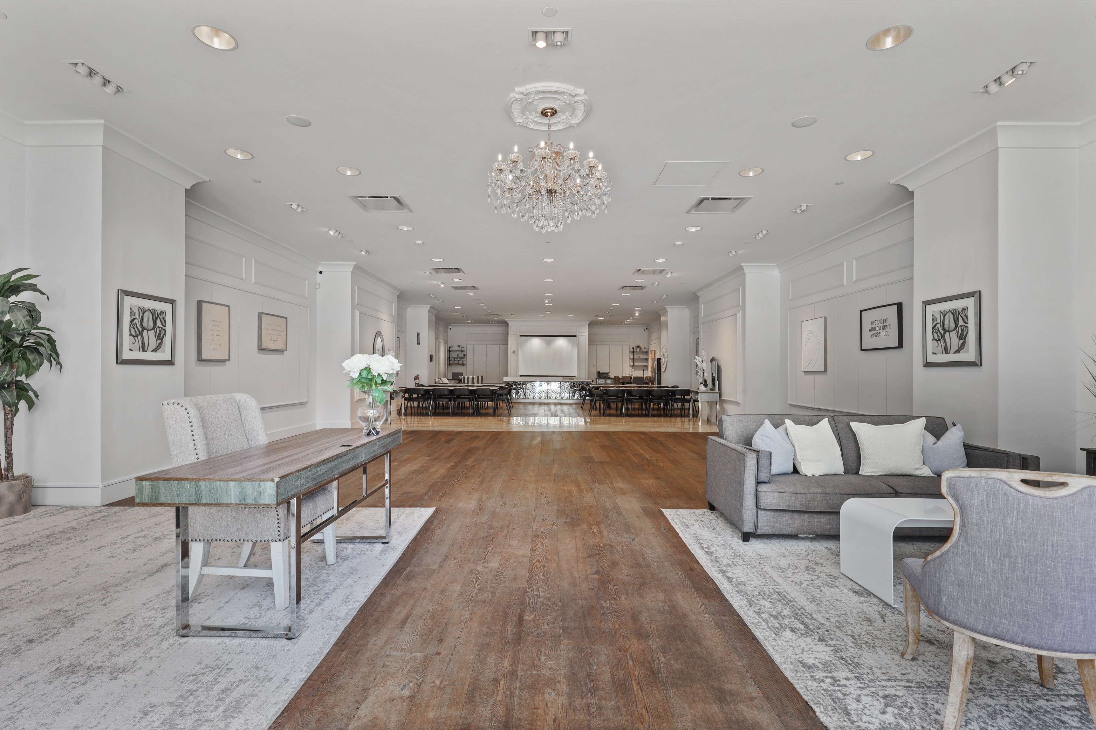 The image shows a spacious, elegantly designed indoor venue featuring hardwood floors, chandeliers, and a mix of seating arrangements, including a sofa and a table.
