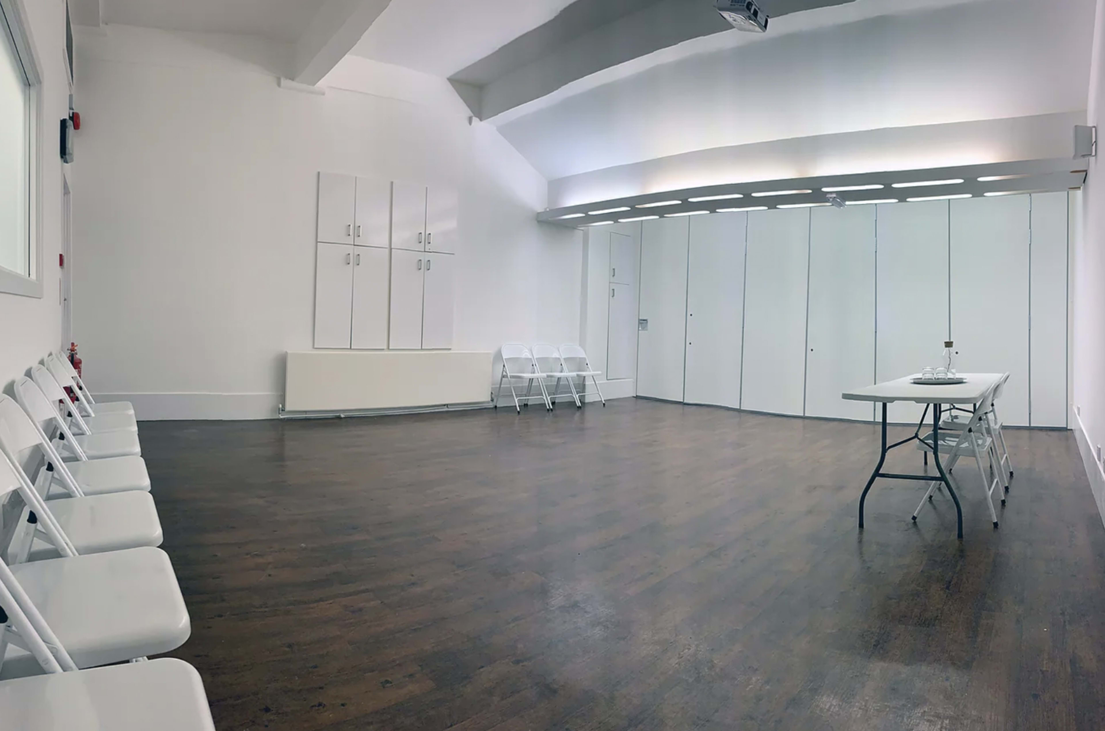 A spacious, empty room with wooden flooring, white walls, a folding table, and several white folding chairs arranged along one side.
