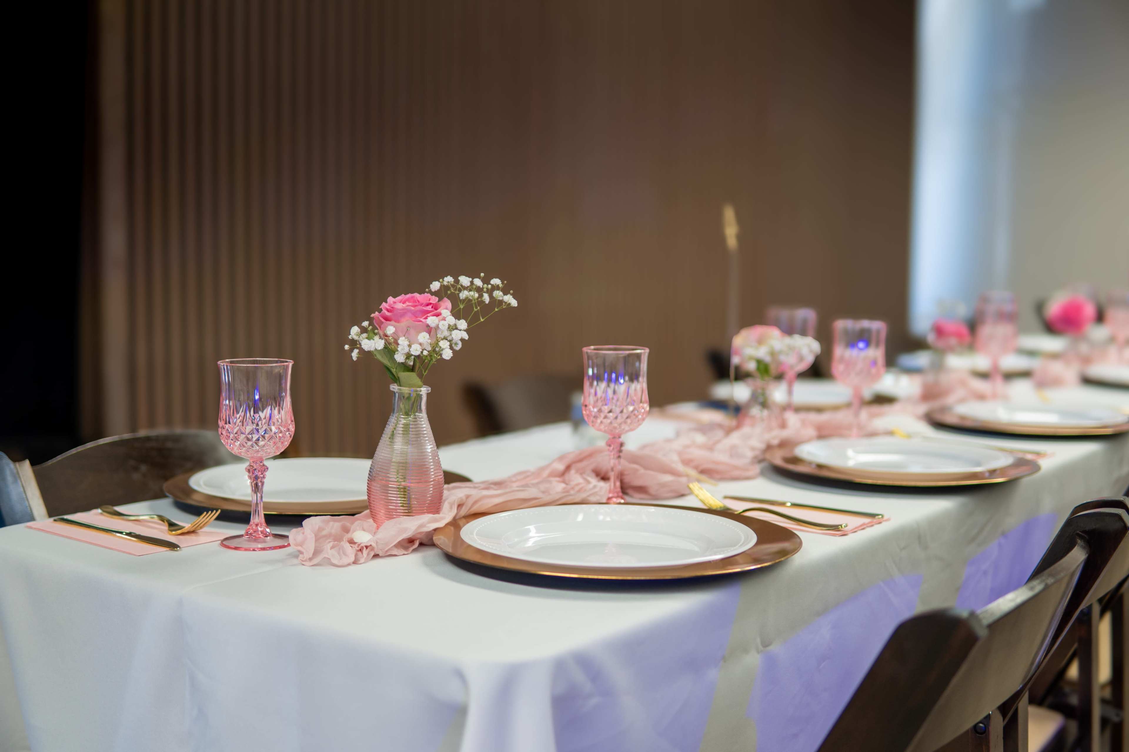 A elegantly set dining table features white plates, gold utensils, pink glassware, and floral centerpieces.