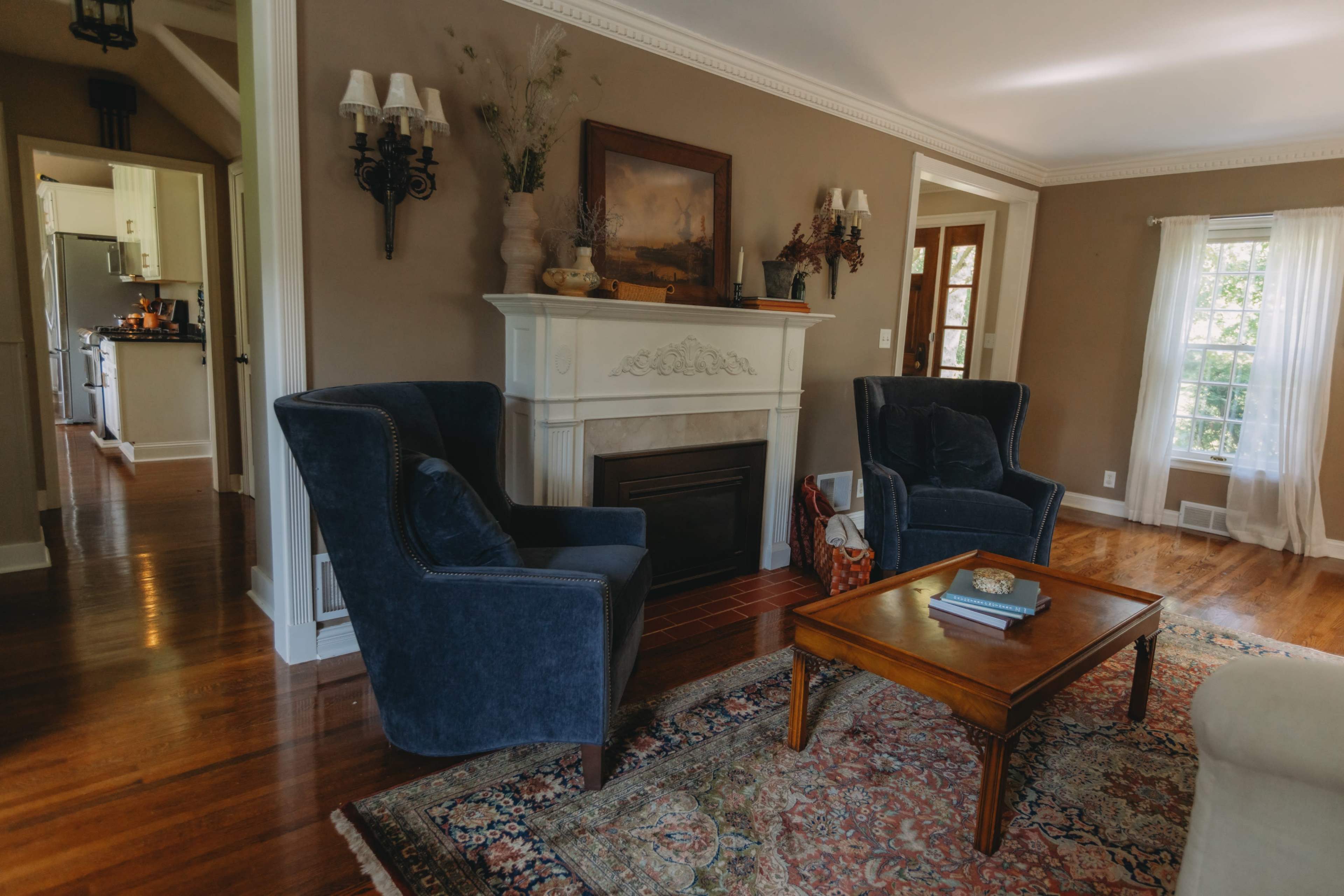 The image shows a cozy living room with two blue armchairs flanking a wooden coffee table, a fireplace with decorative features, and a view of an adjoining kitchen through an open doorway.