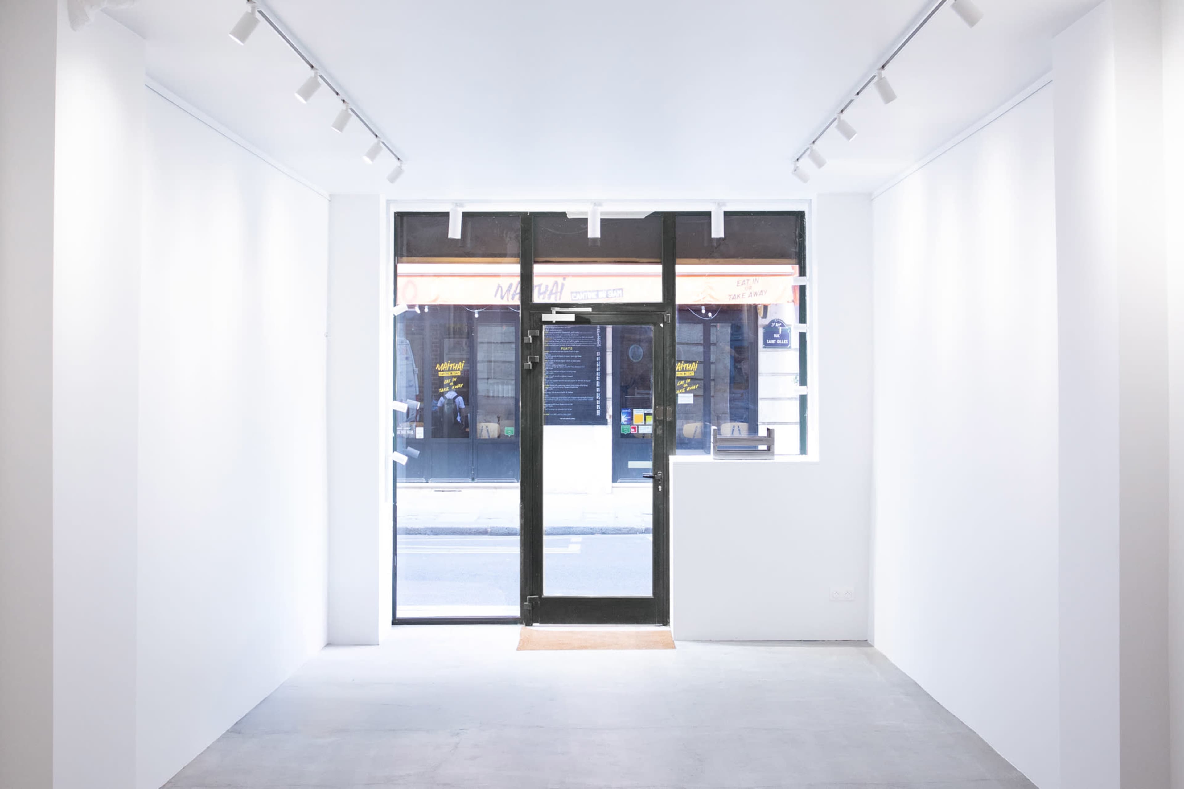 The image shows a minimalistic, empty interior of a store with white walls and a large glass entrance facing an exterior street.