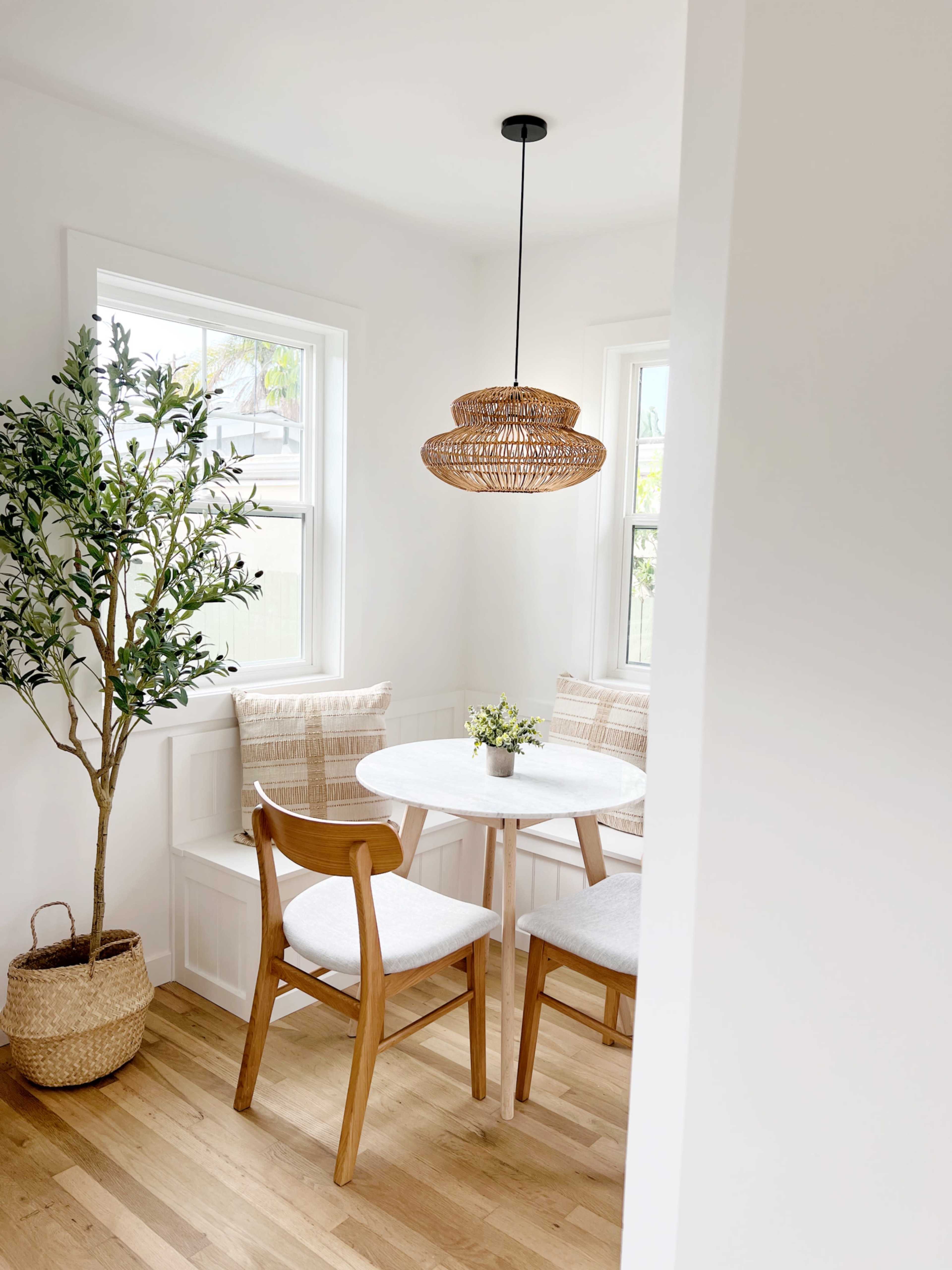 A cozy dining nook features a round table surrounded by wooden chairs, with a hanging light and a small potted plant on the table, beside a tall indoor tree.