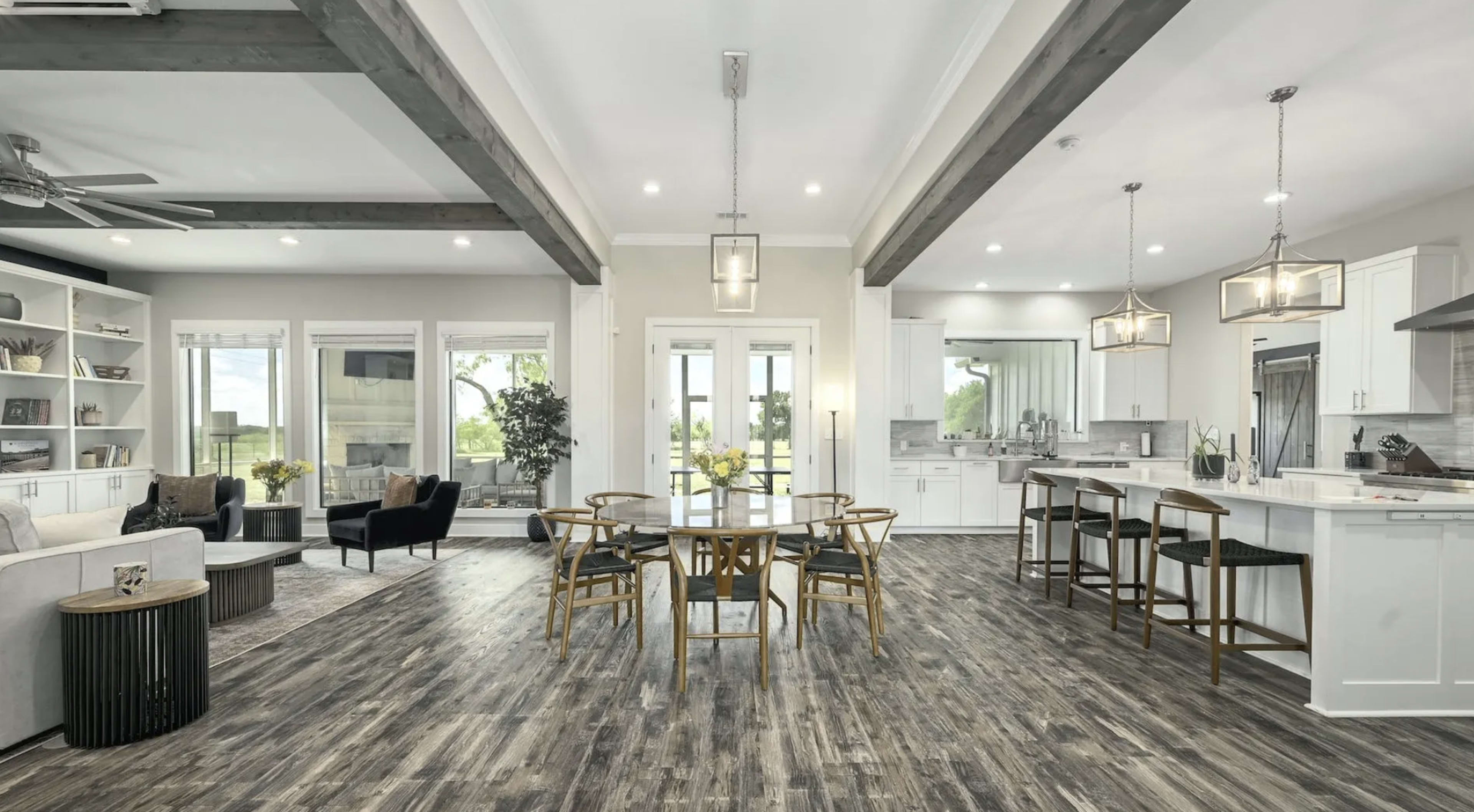 A spacious, modern open-concept living area features a dining table surrounded by chairs, a kitchen in the background, and large windows with views of greenery.
