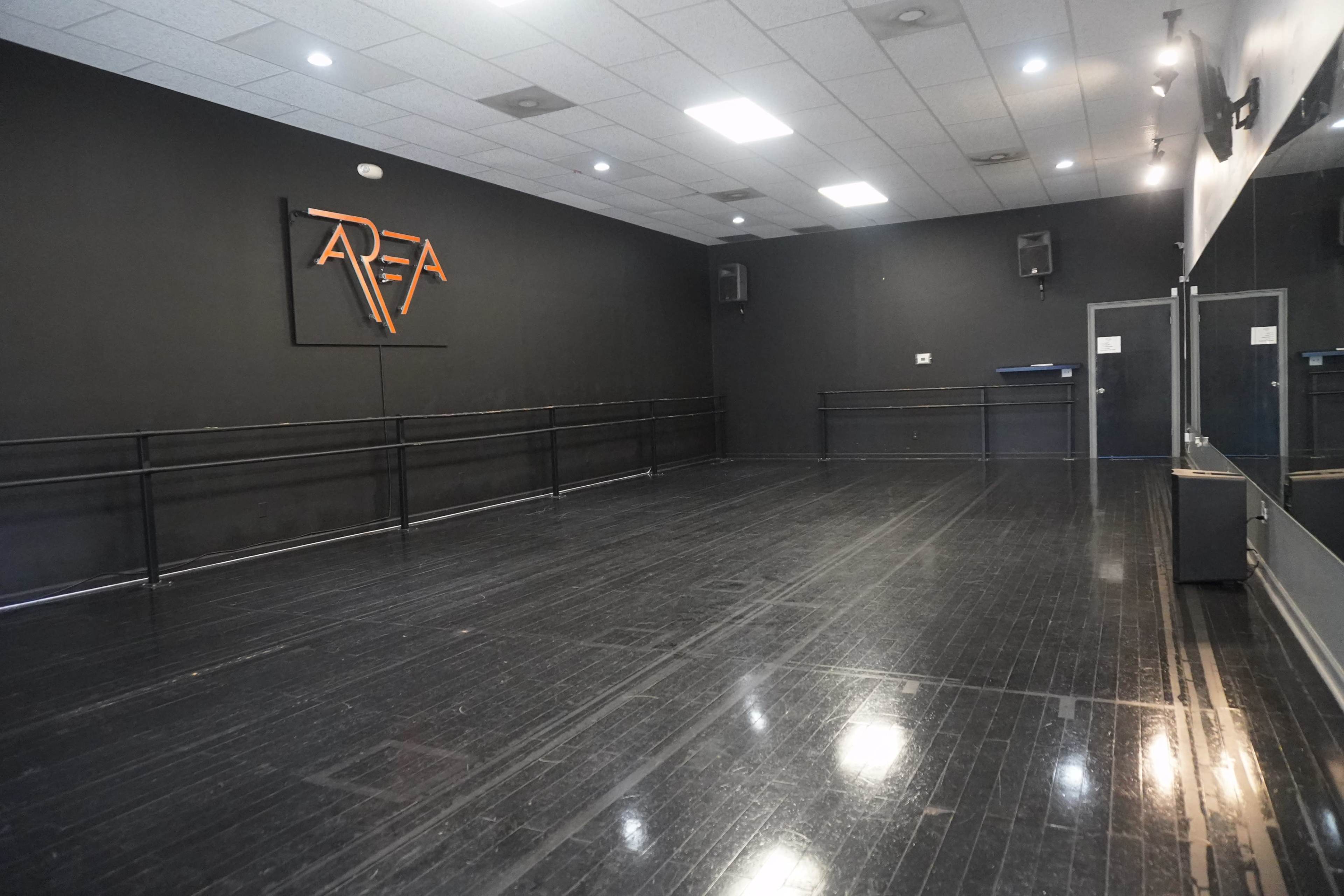A dance studio featuring a black wall with a logo, wooden flooring, and ballet bars along one side.